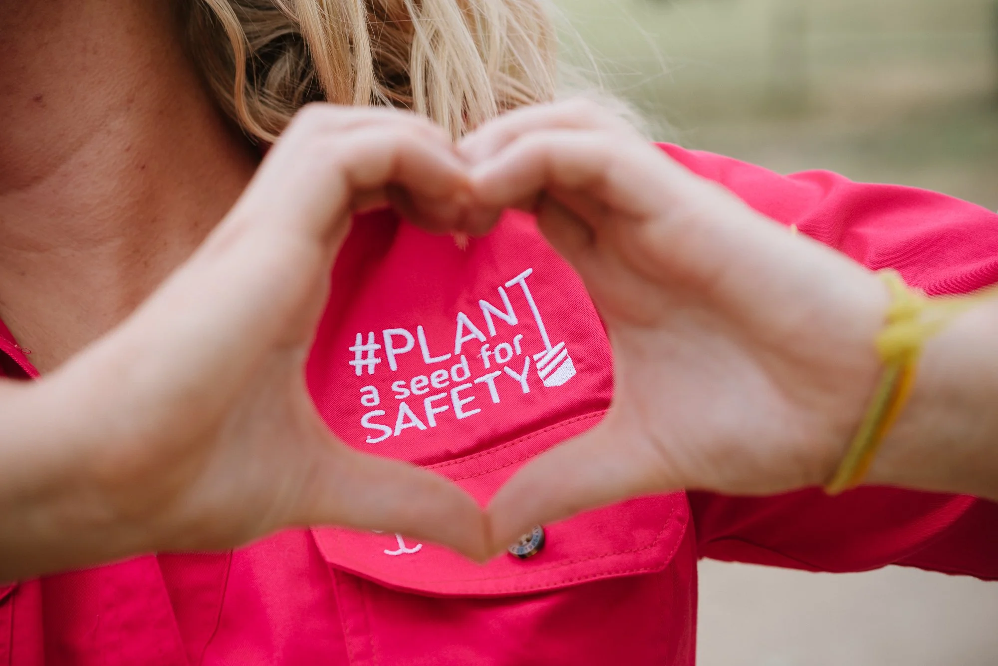 Close-up of a person forming a heart shape with their hands, wearing a pink shirt with a sticker that reads '#PLANT a seed for SAFETY'.