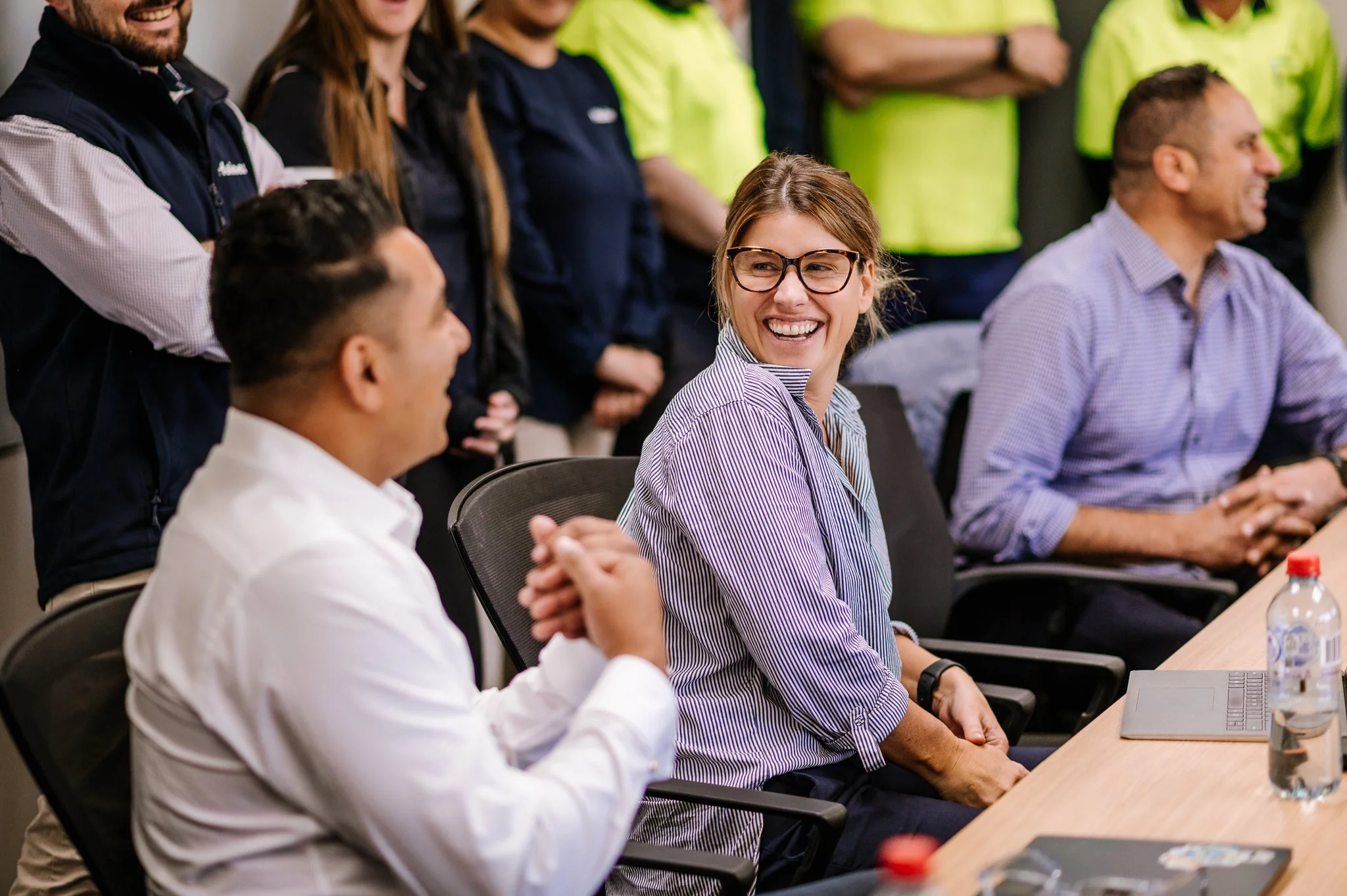 People in a business meeting or office setting, woman with glasses smiling and looking at a man.