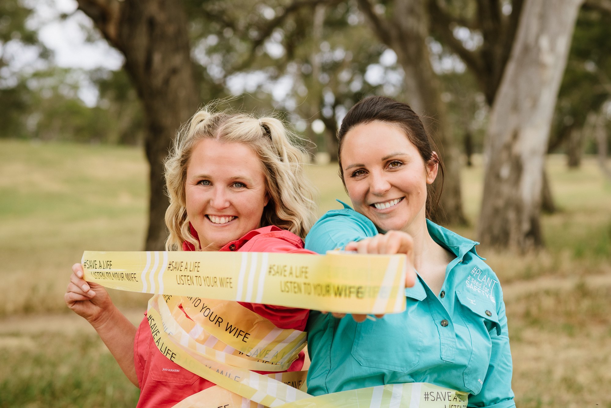 Two women outdoors wrapped in yellow caution tape with message 'save a life listen to your wife', smiling and holding the tape together.