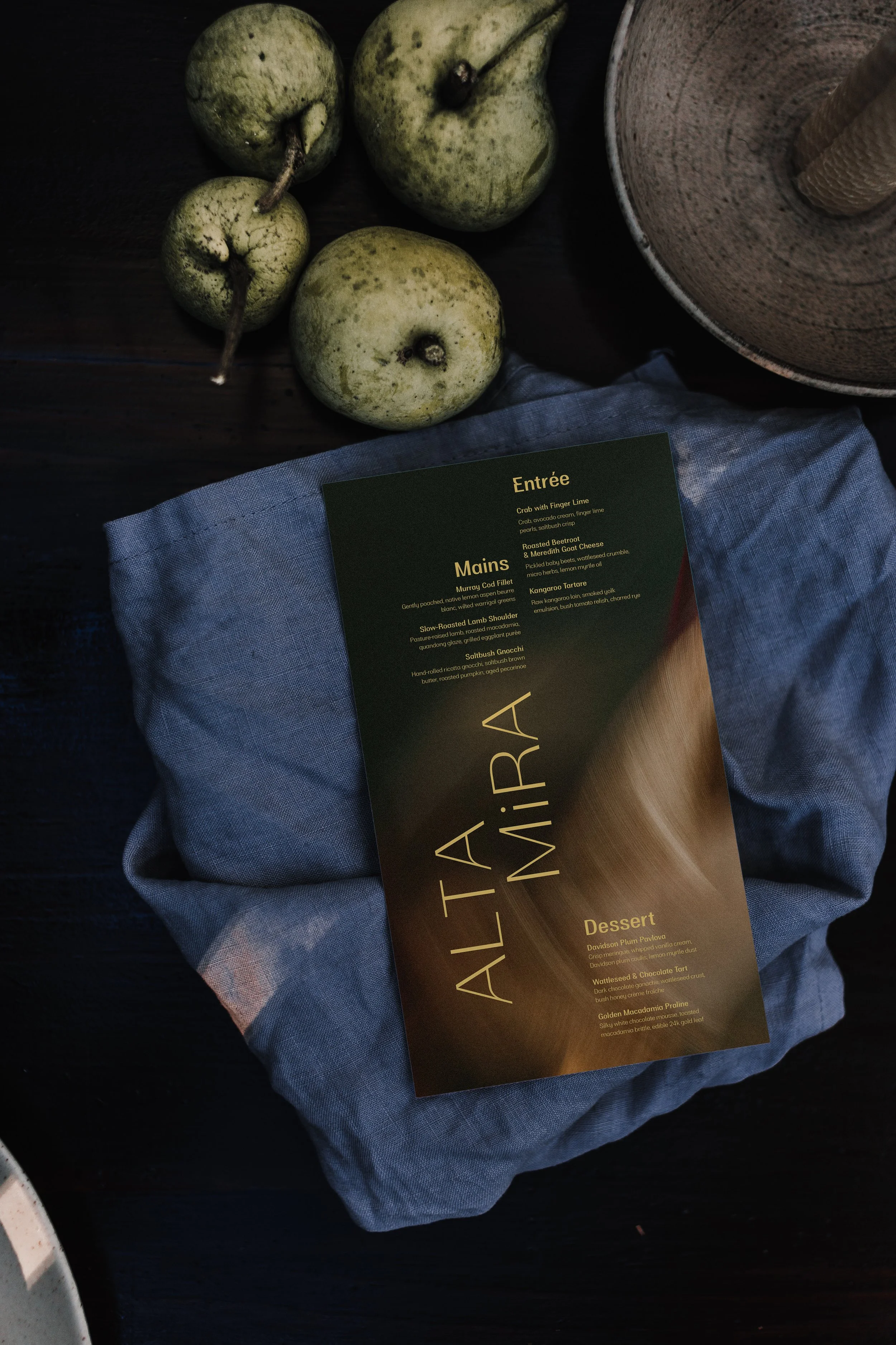 A dark wooden table with three green pears, a blue cloth, a black menu with gold and cream text, and a wooden stand or coiled element in a shallow bowl.