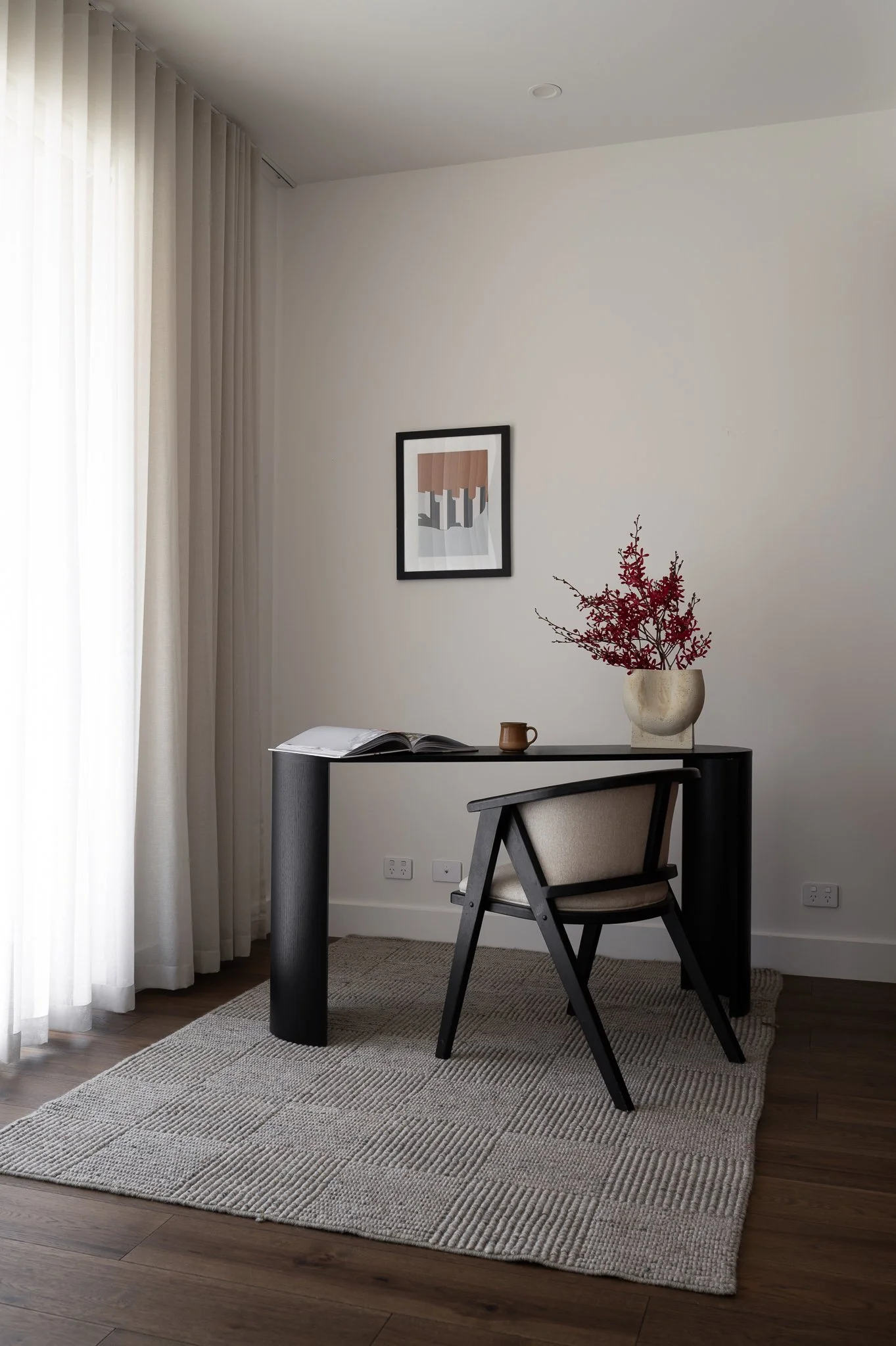Detail view of work nook with a desk and chair. Tones are soft beiges and browns with a vase holding a spray of fine deep red flowers.