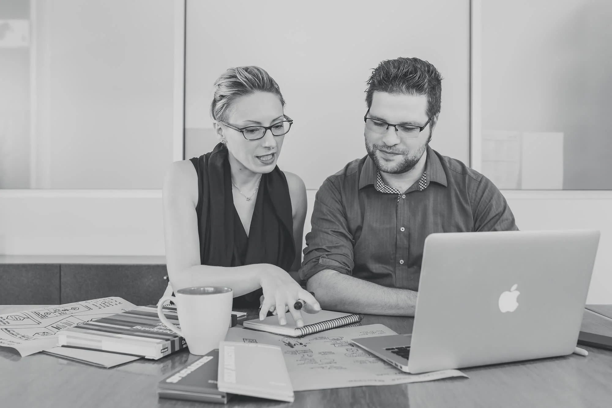 Two colleagues, a woman and a man, collaborating on a project at a table in an office. The woman is gesturing towards a notebook, and both are wearing glasses. There are books, papers, a coffee mug, and a laptop on the table.