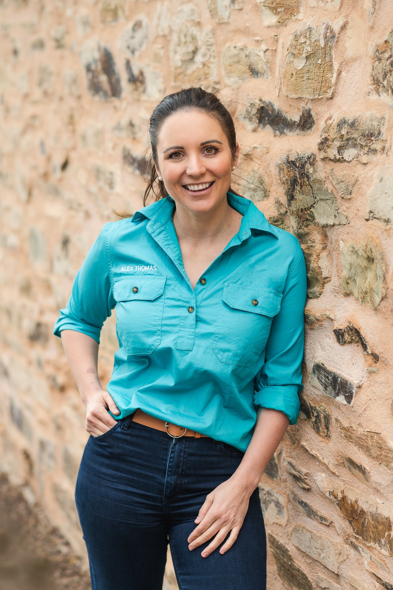 A woman with dark brown hair tied back, wearing a turquoise button-up shirt labeled 'Alex Thomas', standing outdoors with her right hand on her hip, leaning against a textured brick wall, smiling at the camera.