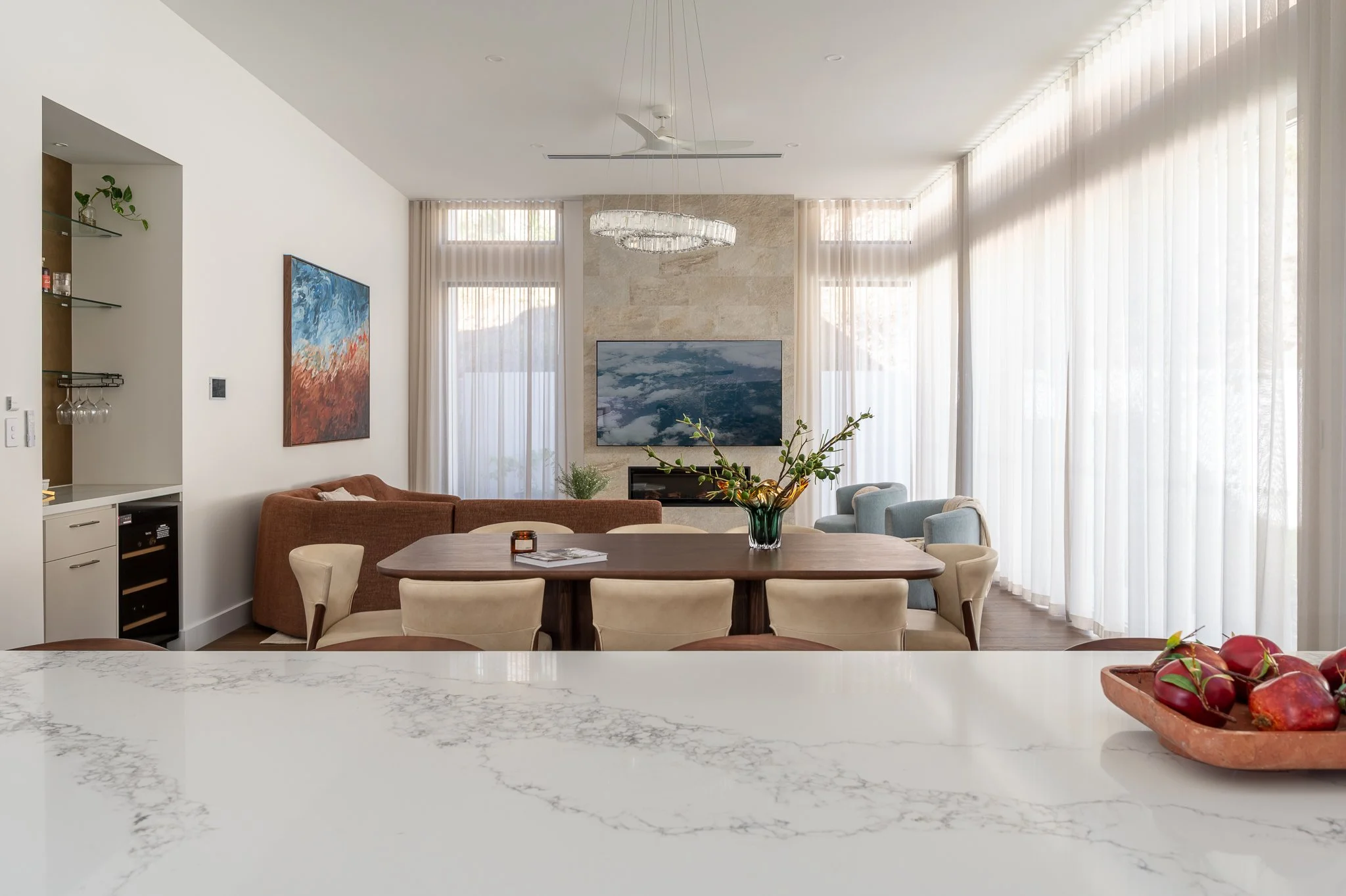 Modern living room with large windows, white curtains, a wall-mounted TV over a fireplace, artwork on the walls, a round dining table with beige chairs, a brown sofa, and a decorative plant on the table.