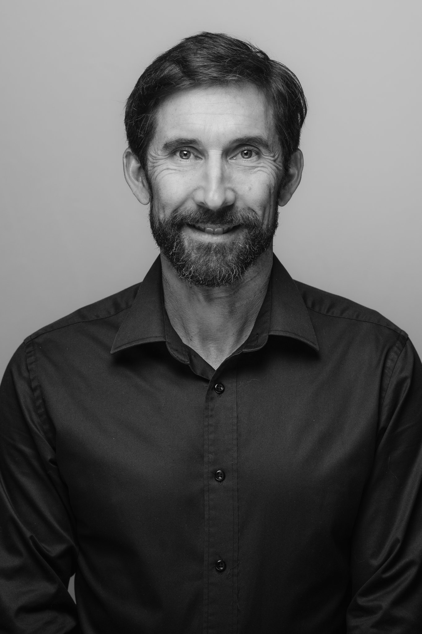 Black and white portrait of a middle-aged man with dark hair and a beard, wearing a black button-up shirt, smiling.