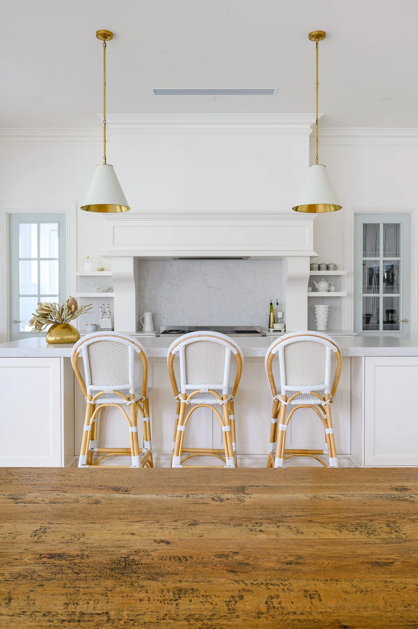 Modern white kitchen with marble backsplash, three rattan barstools with white woven seats, and gold pendant lights hanging from a white ceiling.