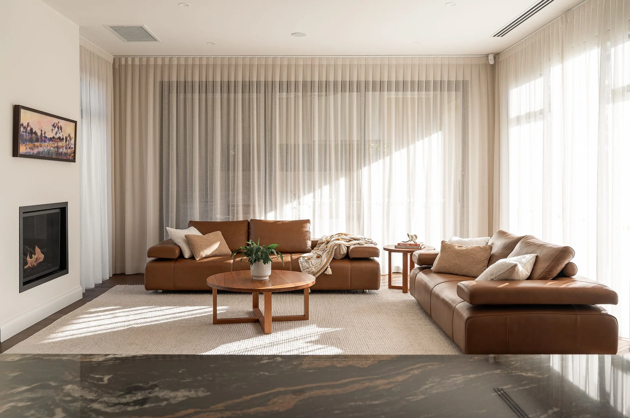 Living room with two brown leather sofas, a round wooden coffee table with a plant, side table with decorations, beige carpet, white curtains, and a painting above the fireplace.