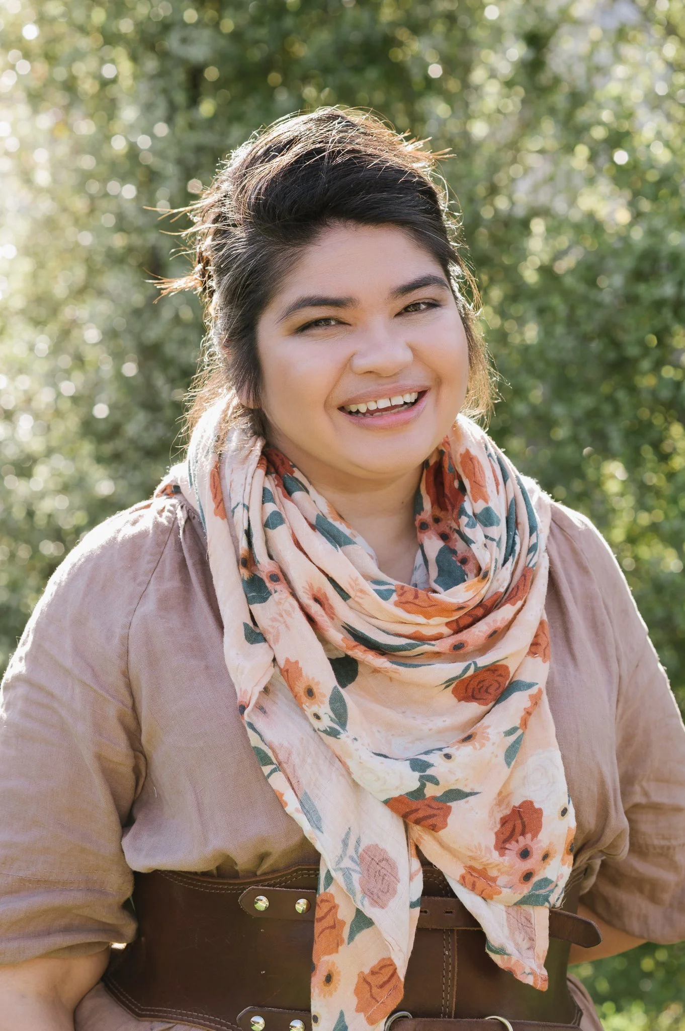 A woman with dark hair smiling outdoors with a blurred green background. She is wearing a beige dress, a brown belt, and a light-colored scarf with orange and green floral patterns.