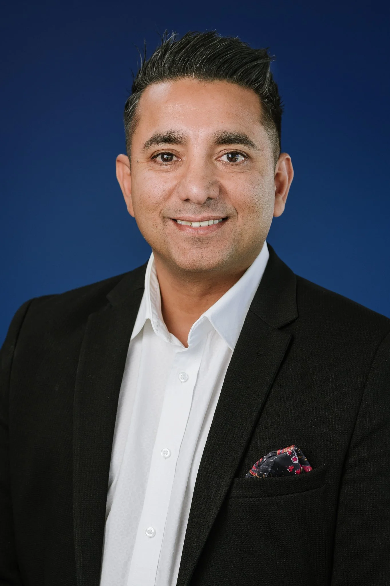 Professional headshot of a man wearing a black blazer, white shirt with a pocket square, smiling against a dark blue background.