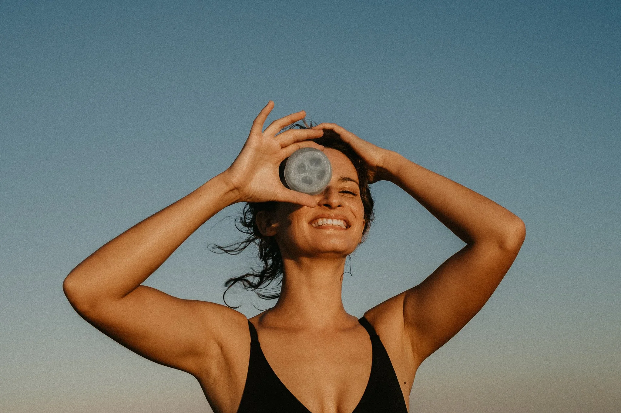 Smiling woman in a black tank top holds a loofah soap over one eye against a clear blue sky.
