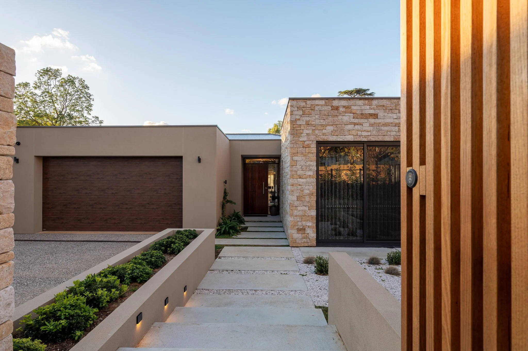 Modern house entrance with stone, wood, and concrete elements, pathway with small plants, and clear sky in background.