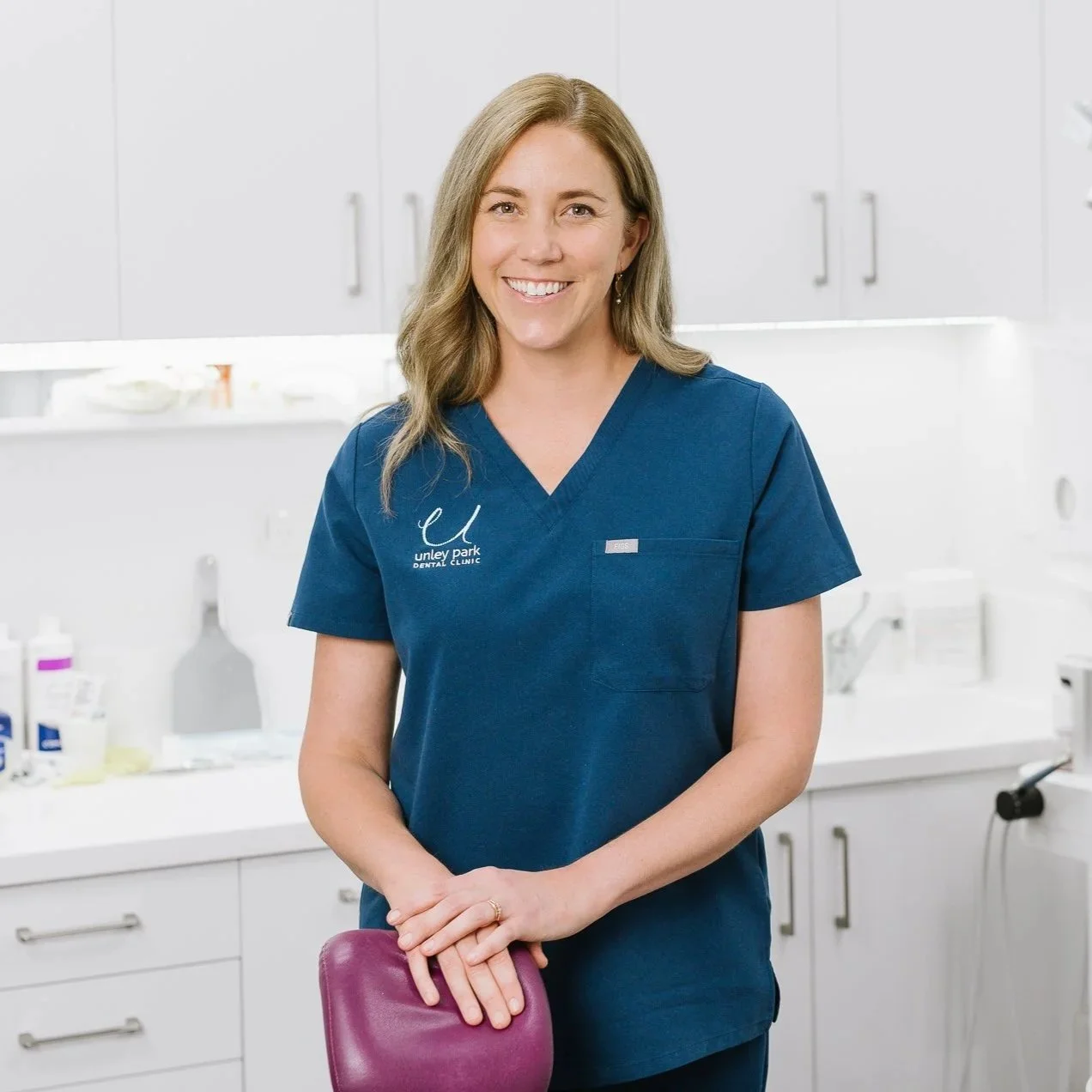 A smiling woman in a blue dental uniform standing in a dental clinic. She is leaning on a purple dental chair, with dental equipment and supplies in the background.