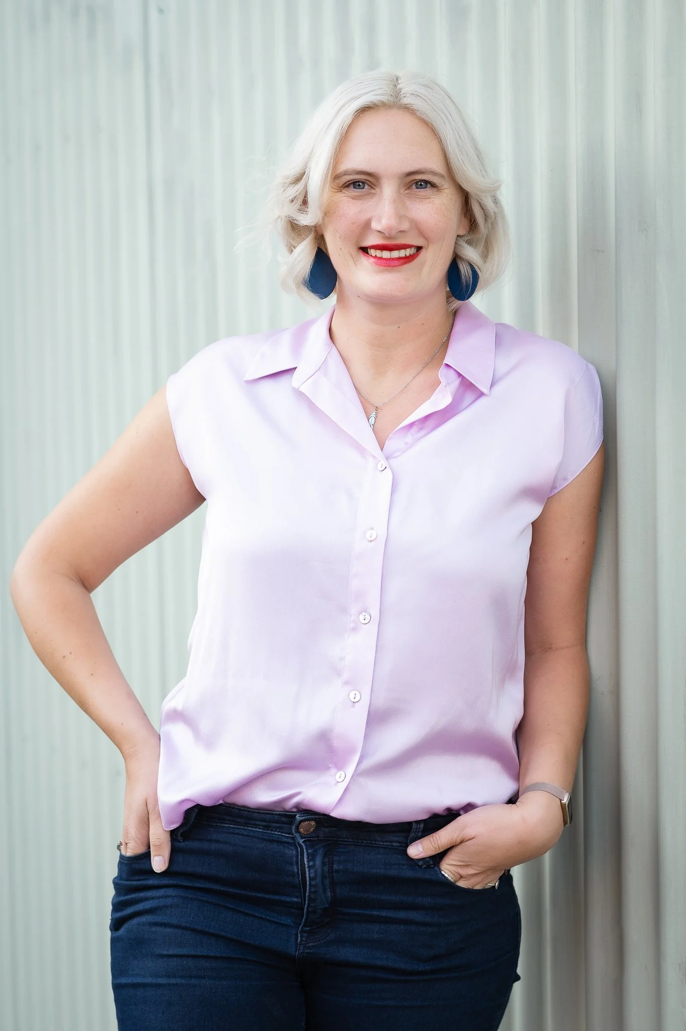 Woman with short blonde hair, wearing a light purple blouse, dark jeans, red lipstick, blue earrings, and a necklace, standing outdoors against a light-colored vertical panel wall, smiling with one hand in her pocket.