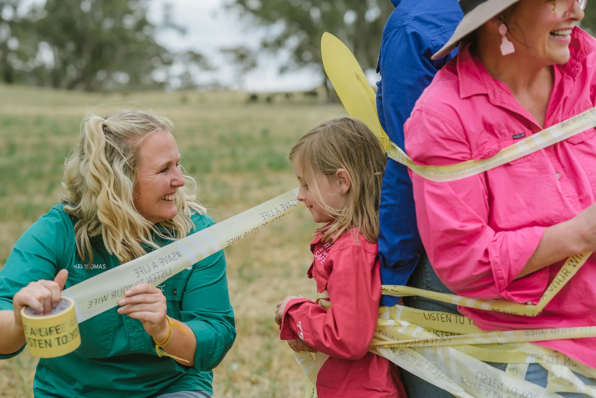 A woman, smiling and talking to a young girl wrapped in yellow caution tape, with another woman nearby, outdoors in a grassy field.