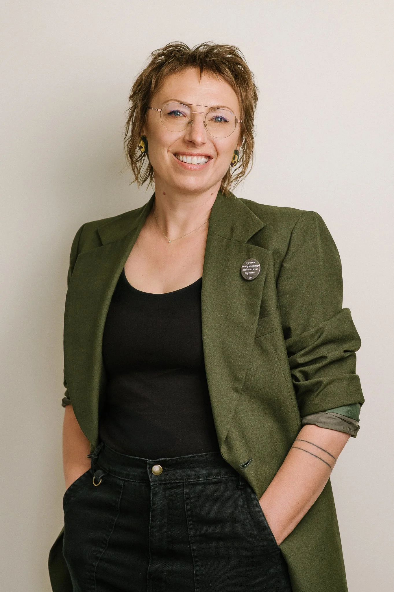 A woman wearing glasses, a green blazer with a pin, and a black shirt, smiling with her hands in her pockets against a plain background.