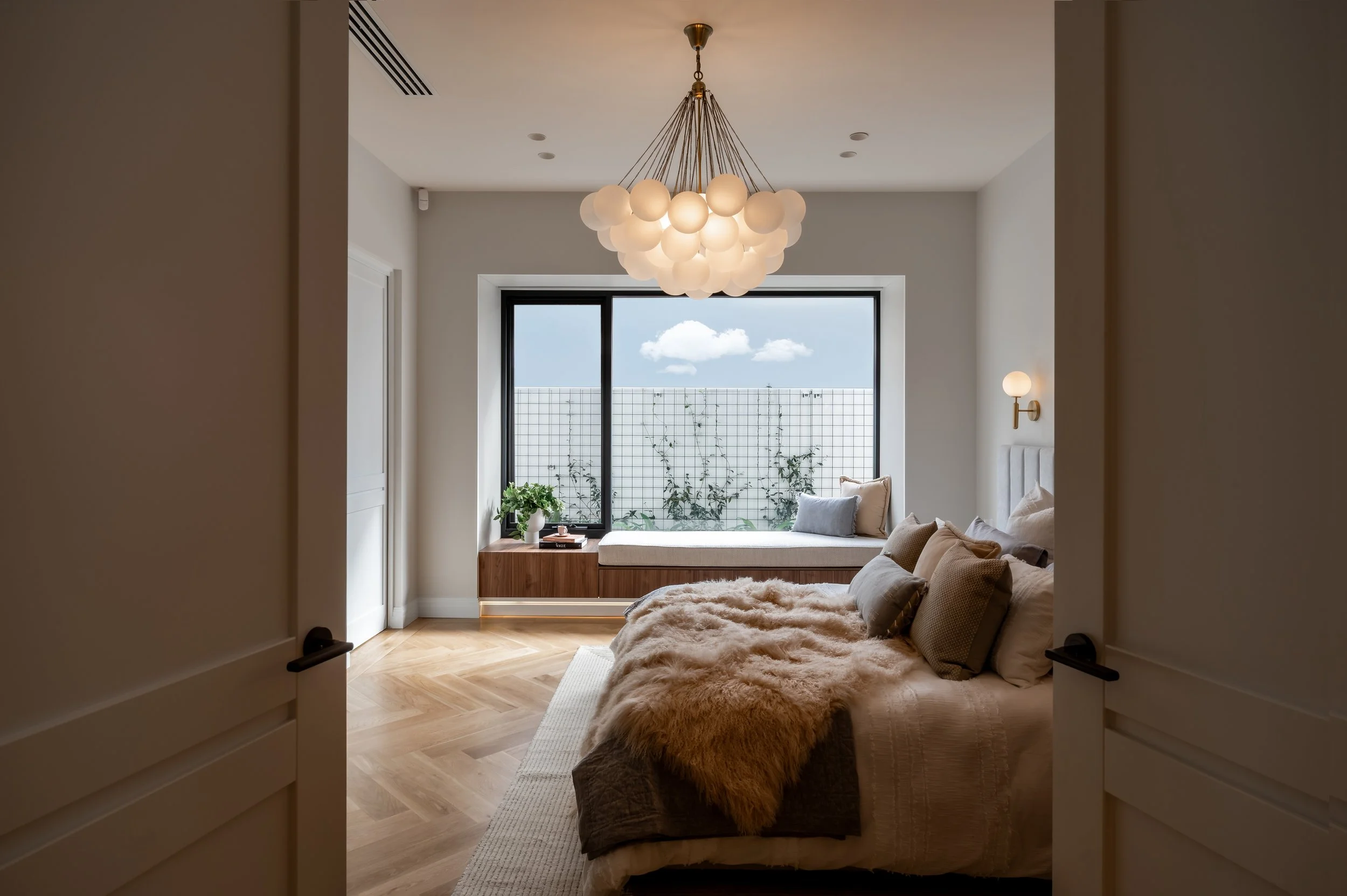 Modern bedroom with a large window, a cozy seating area, and a contemporary chandelier. The room features a bed with pillows and a plush rug on light wooden flooring.