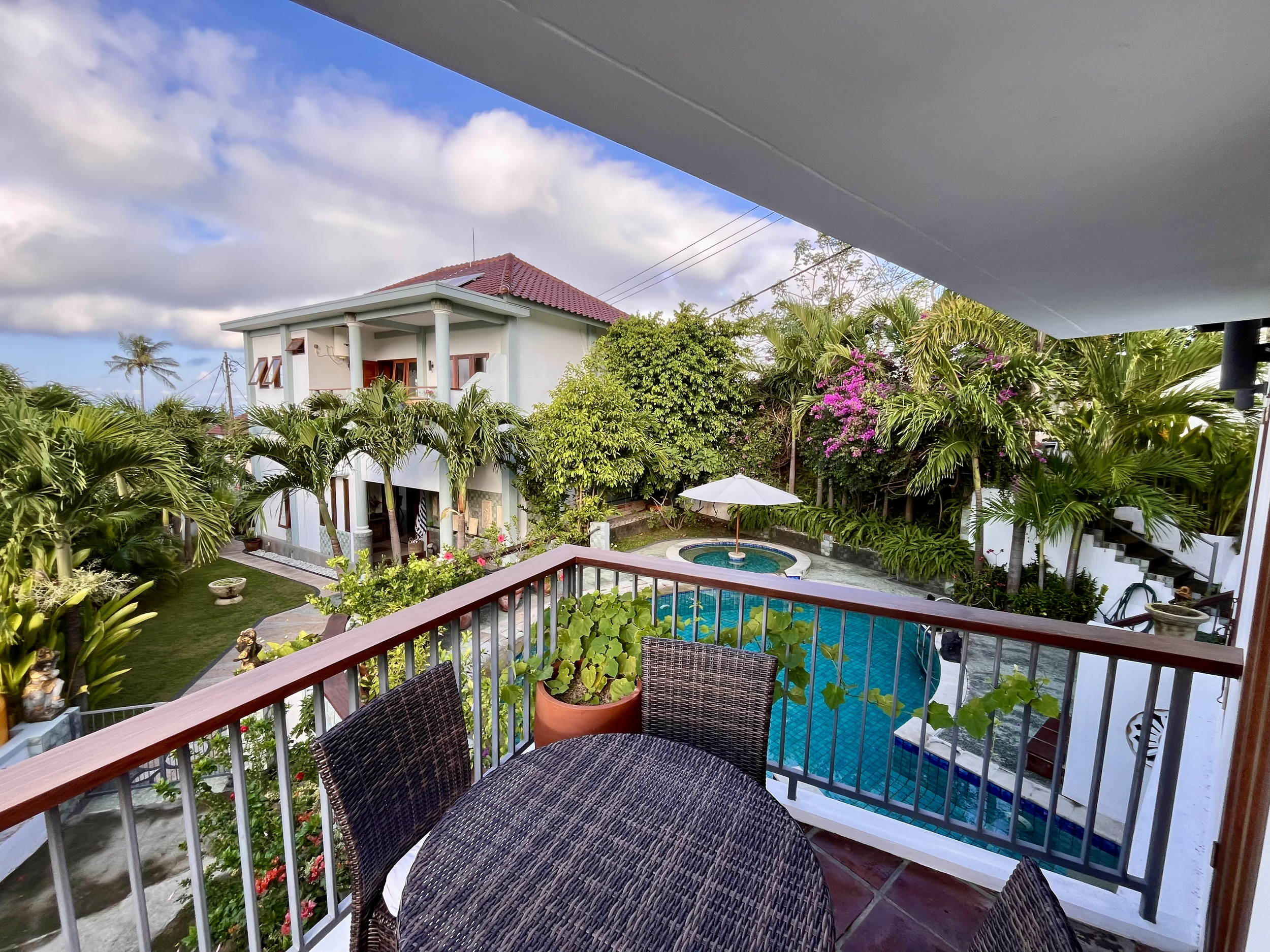 View from a balcony overlooking the tropical backyard with a pool, palm trees, colorful flowers, and main house with a red-tiled roof.
