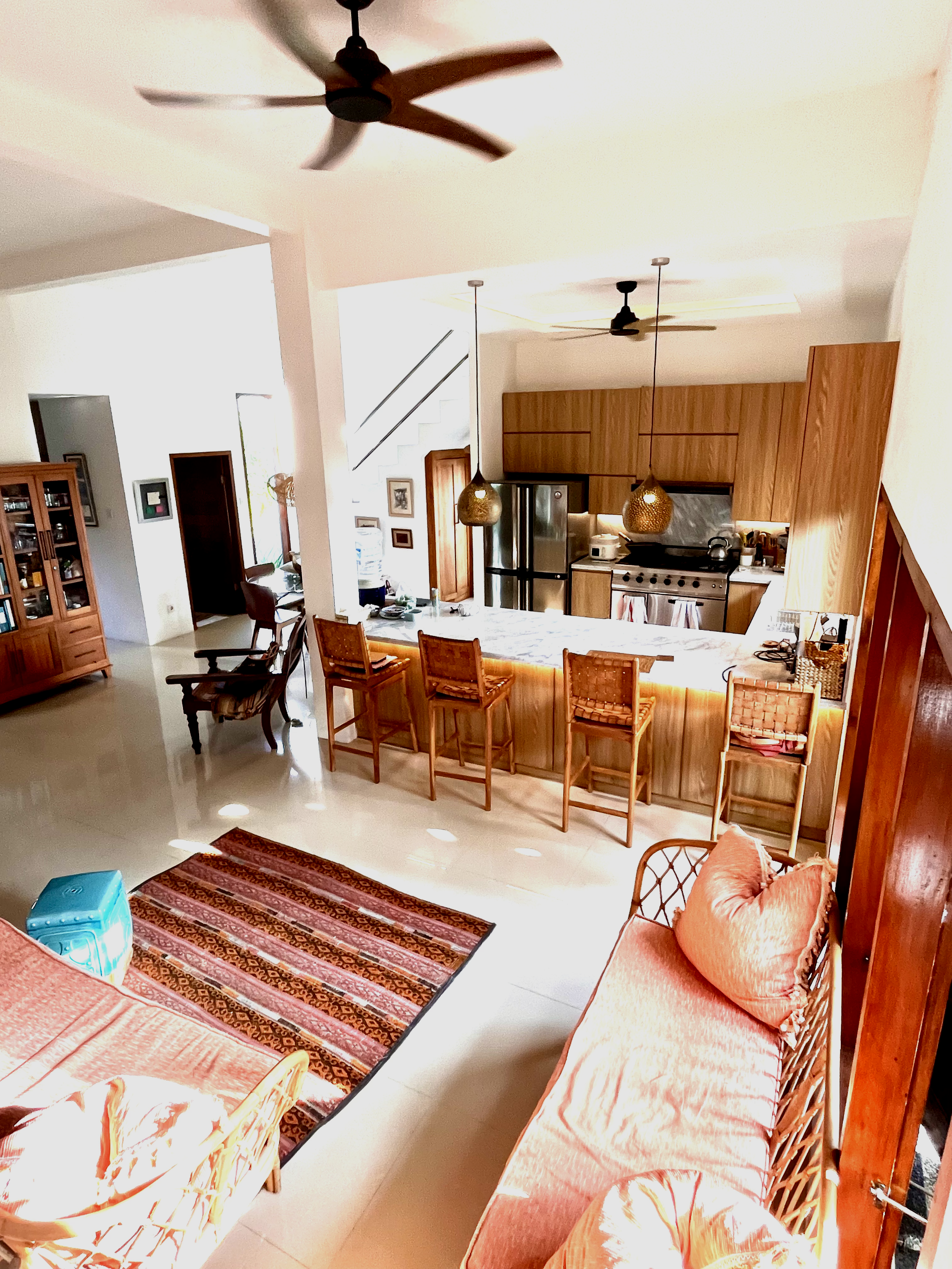 View of a living room with a kitchen area, barstools, wooden cabinets, stainless steel appliances, ceiling fans, and a colorful rug.