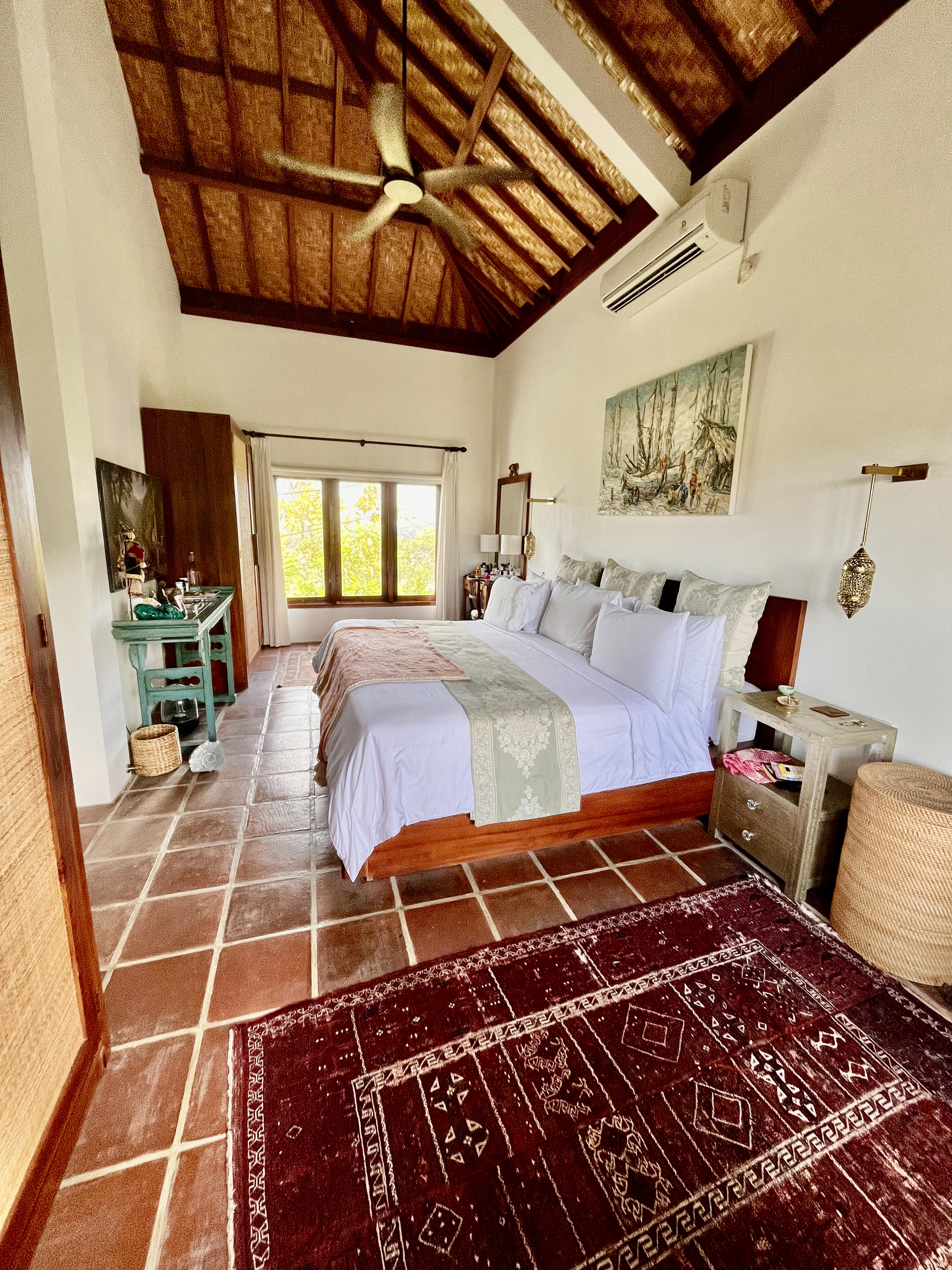 A bedroom with a high wooden ceiling, a ceiling fan, a window with lush greenery outside, a large bed with white linens and pillows, a side table, a wall-mounted air conditioner, and a painting above the bed. The room has tile flooring and a red patt