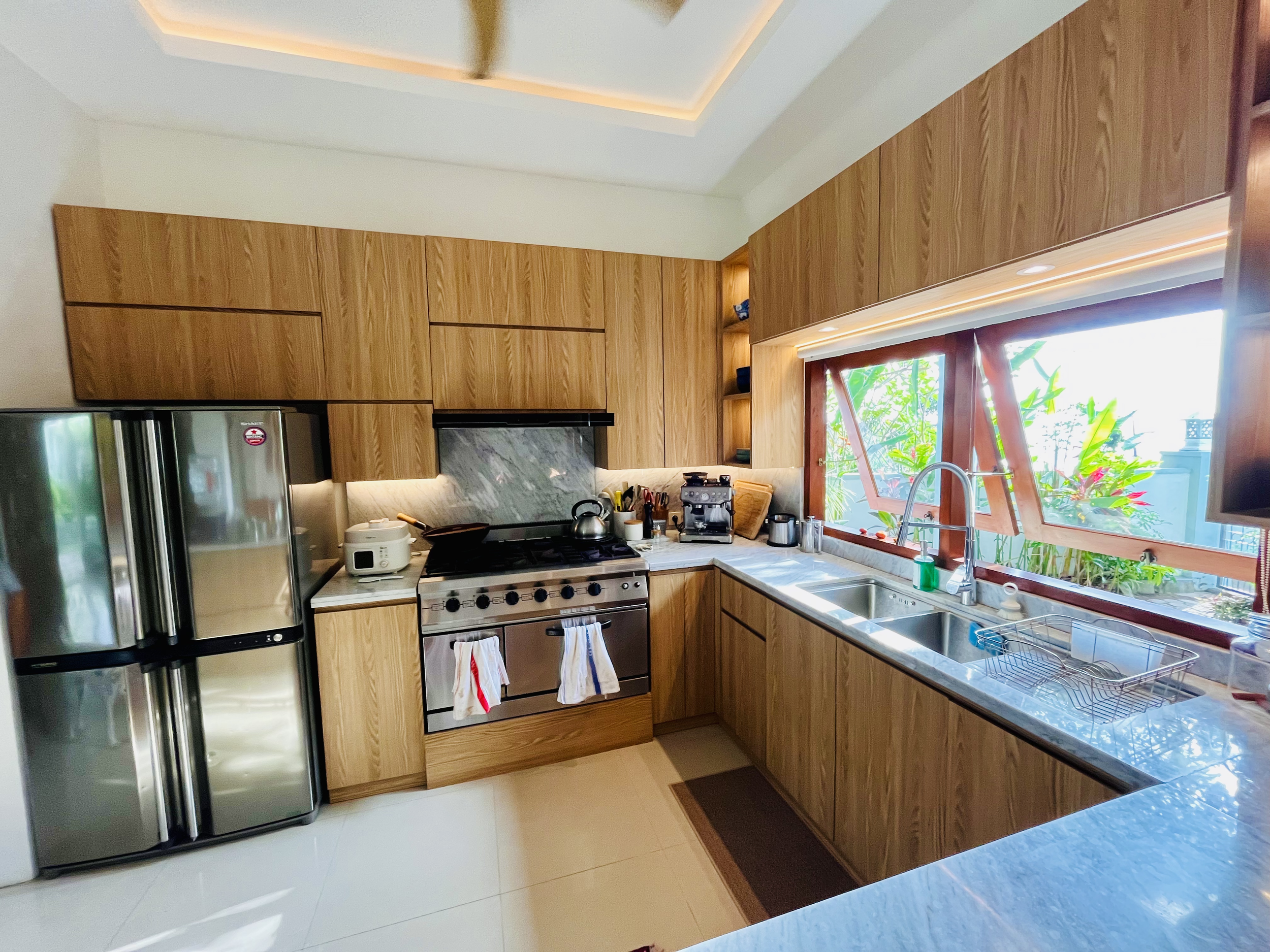 Modern kitchen with wooden cabinets, stainless steel refrigerator, and a large window overlooking Senggigi Bay and Bali's Mount Agung through the lush back garden.