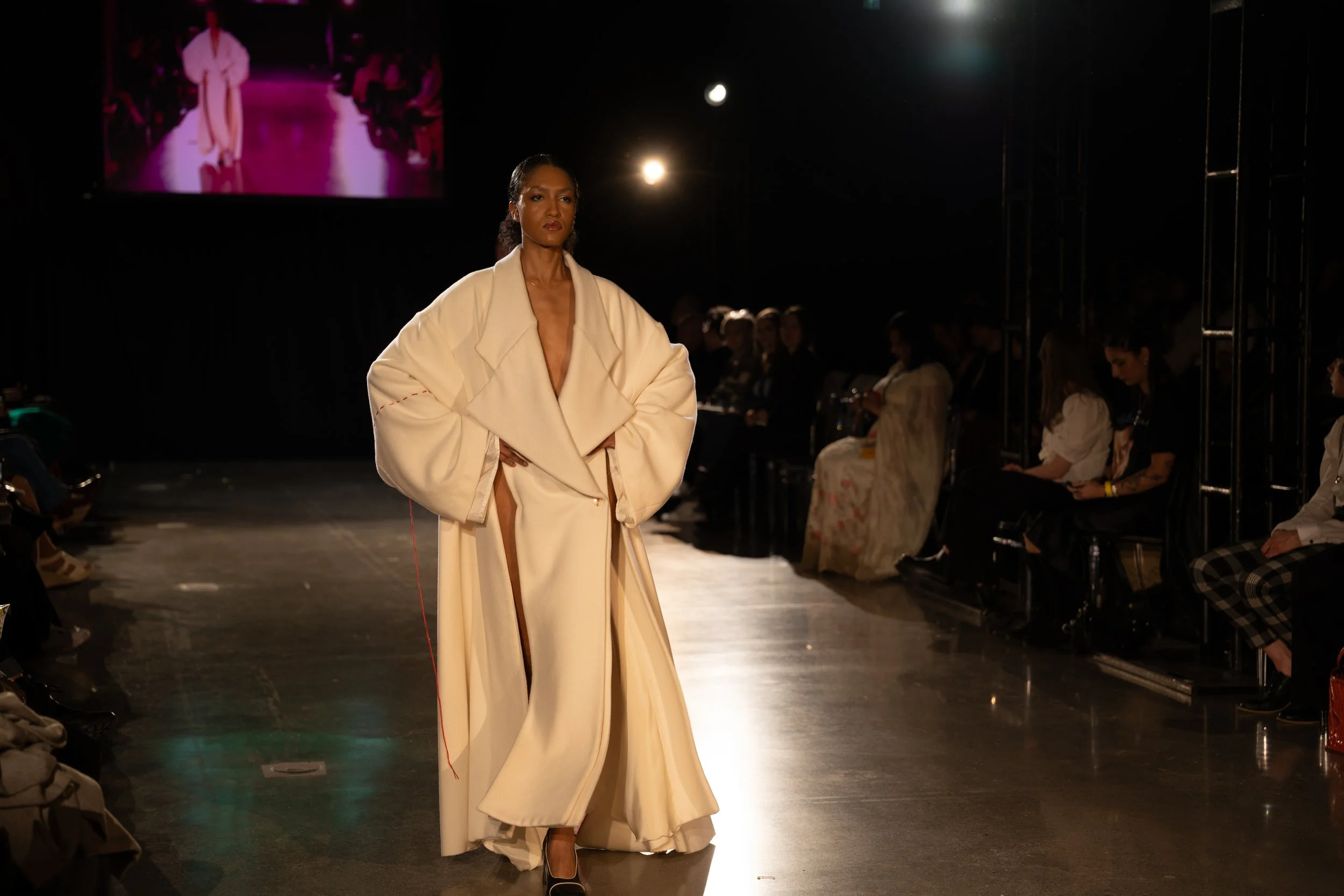 Une femme marche sur un podium lors d'un défilé de mode, portant une tenue élégante beige avec un large col et des manches bouffantes, dans un environnement sombre avec une audience assise sur les côtés.
