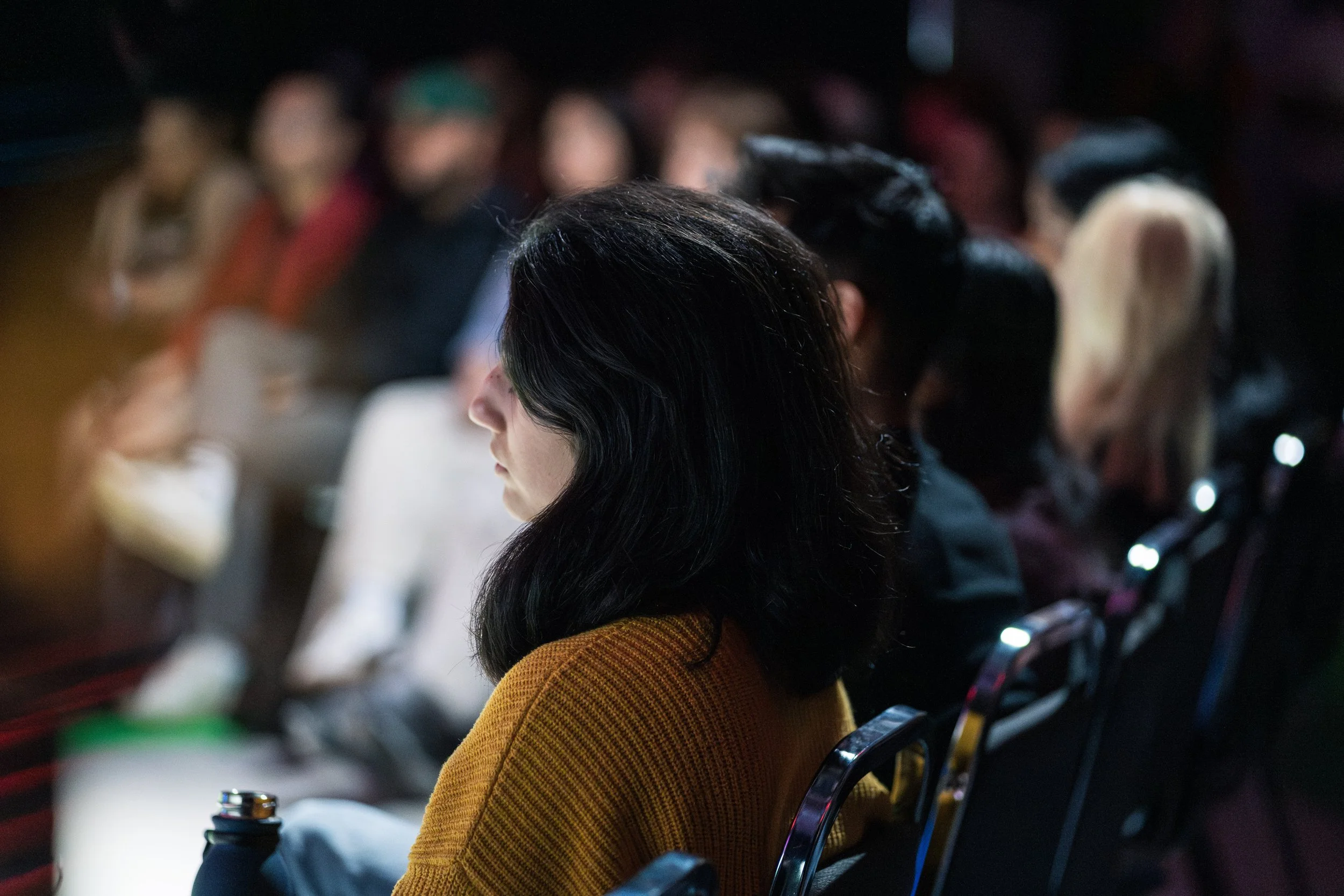 Une femme aux cheveux noirs portant un pull jaune assise parmi un groupe de personnes lors d'une conférence ou d’un événement en intérieur, en prise de vue de profil.