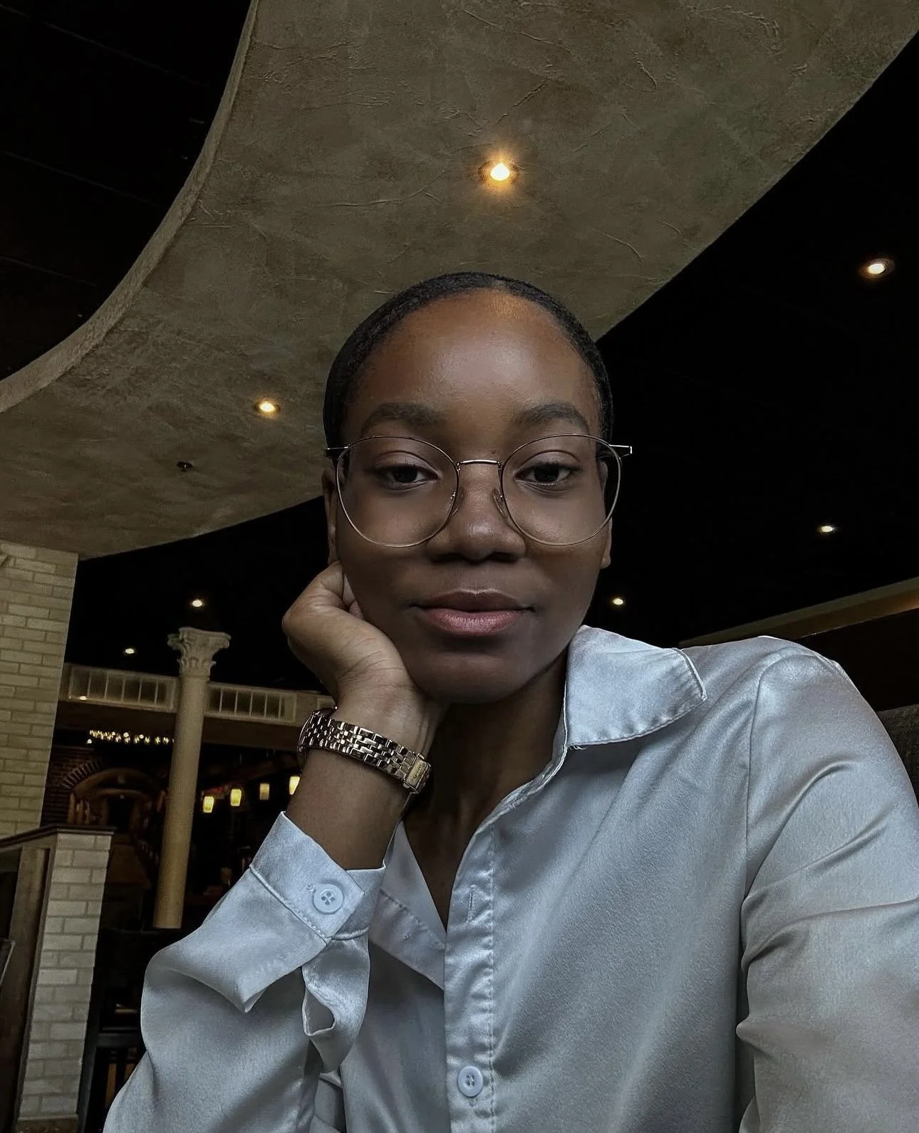 Portrait d'une femme portant des lunettes, vêtue d'une chemise en satin, dans un intérieur avec plafond en béton et lumières encastrées.