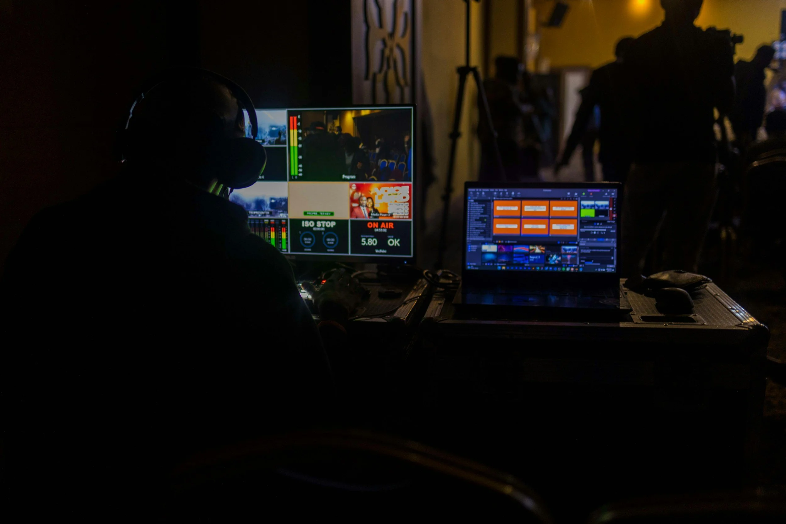 Darkroom with video editing and live streaming equipment, featuring multiple monitors displaying video feeds, audio levels, and editing software, with a silhouette of a person wearing headphones working at the desk.