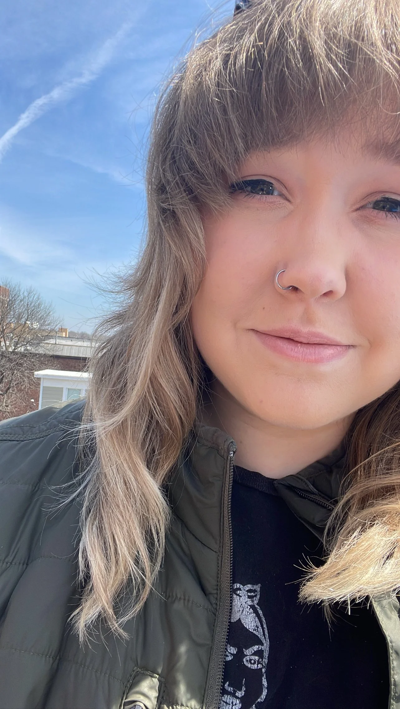 Close-up of a young woman outdoors smiling, wearing a dark jacket and black shirt with a graphic print, with a blue sky and some trees in the background.