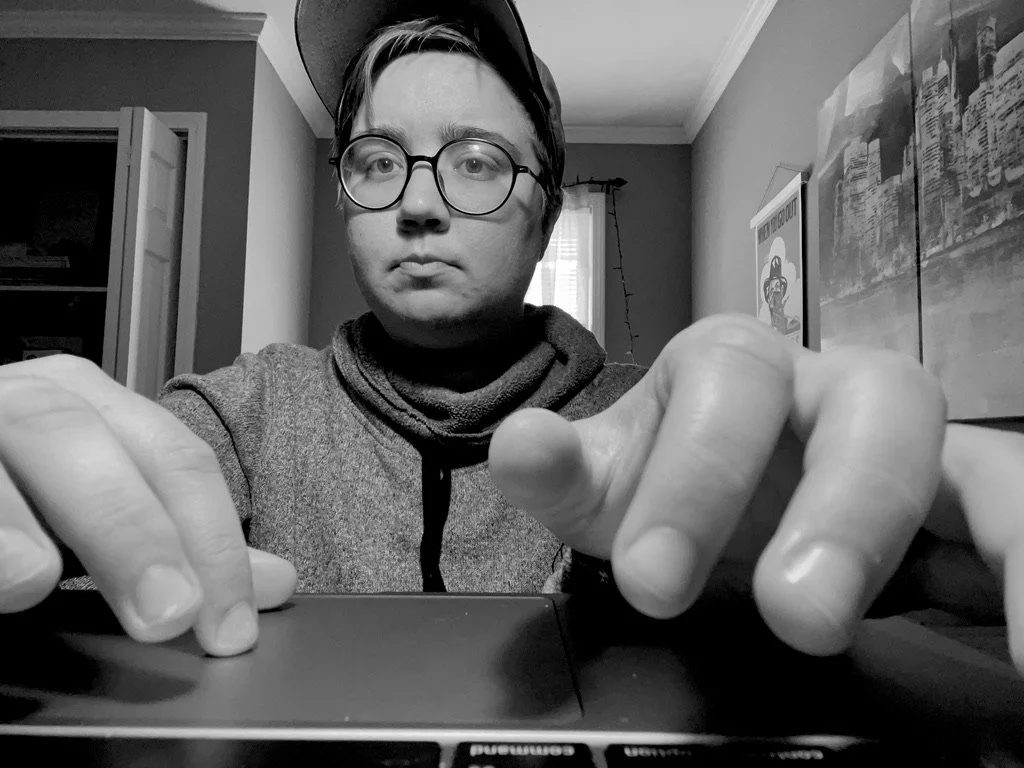 A young person with glasses and a cap taking a selfie, with their hand on a laptop keyboard in a room with a window, posters, and a door in the background.