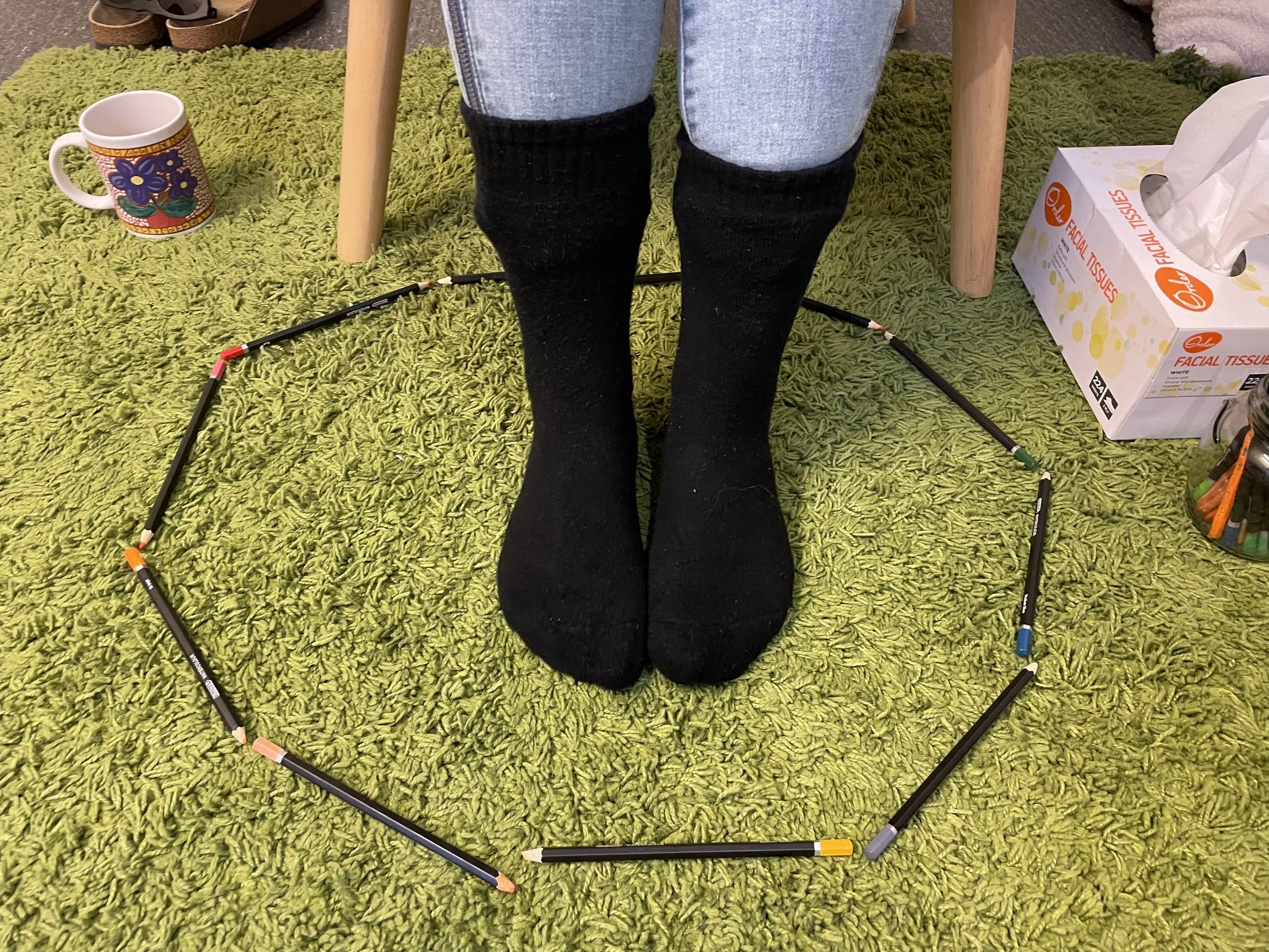 Person wearing black socks and jeans standing on a green shag carpet, surrounded by a circle of colored pencils, with a mug on the left and facial tissue box on the right.
