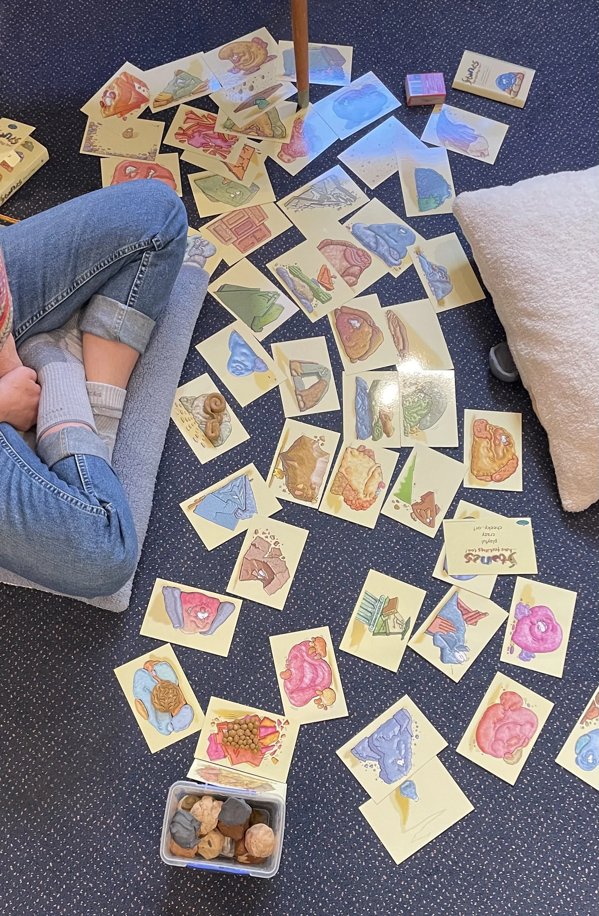 A child sitting on a gray cushion on a dark blue carpet with small colorful dots, playing with a set of illustrated matching cards. The cards feature various colored rocks or minerals. A small plastic container filled with different colored rocks or minerals is next to the child. There is a fluffy, beige pillow on the right side of the image.