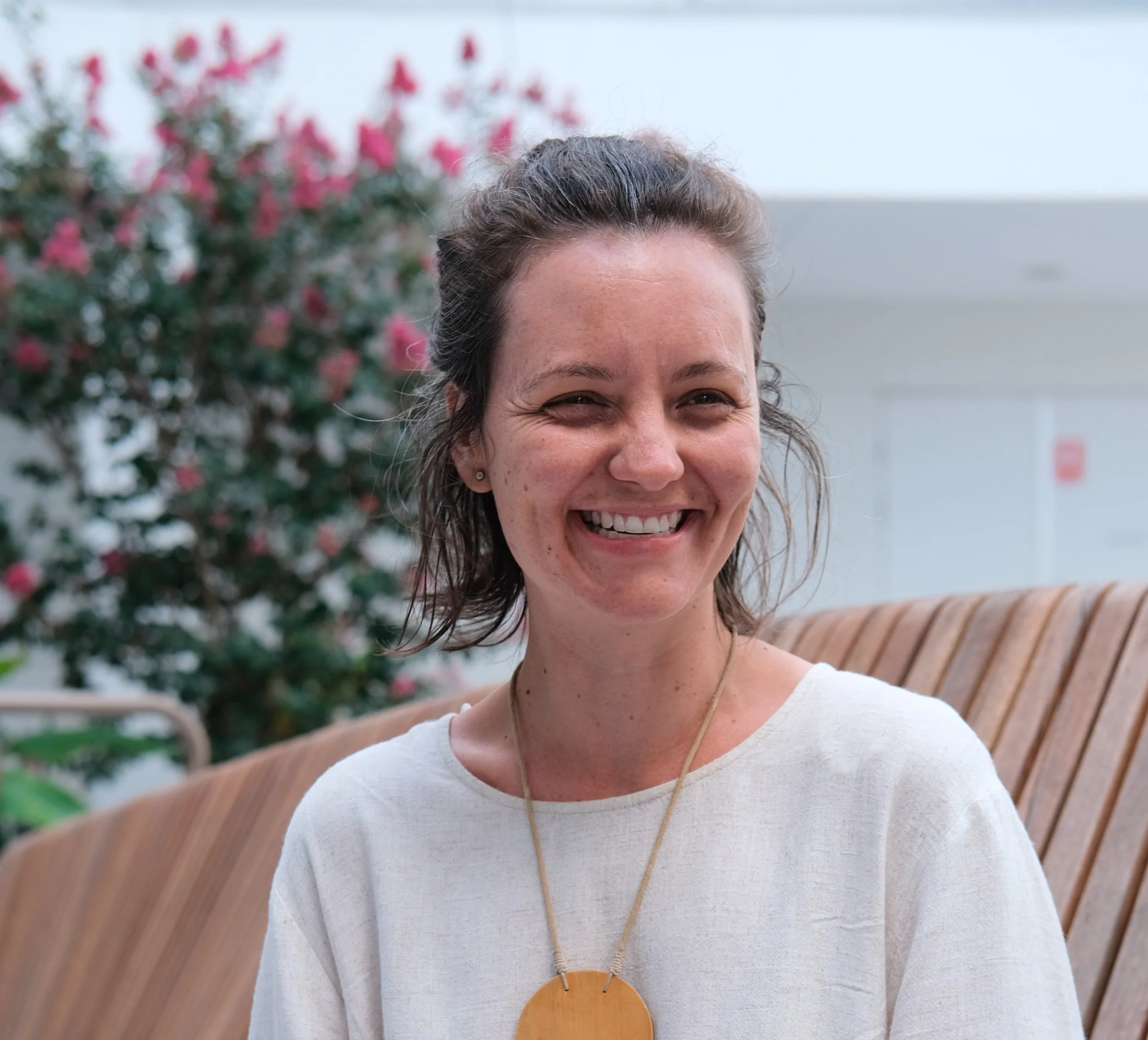 A woman with short brown hair smiling, sitting on a wooden bench outdoors, with pink flowering bush and a white wall in the background.