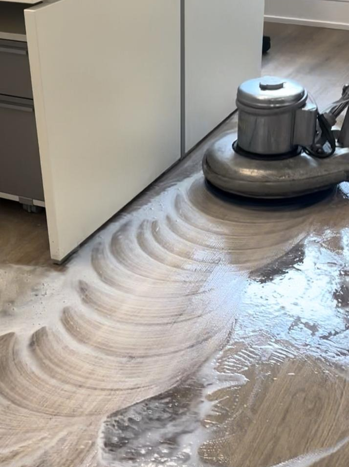 A floor cleaning machine is polished over a wooden floor, creating swirling patterns with soap and water.