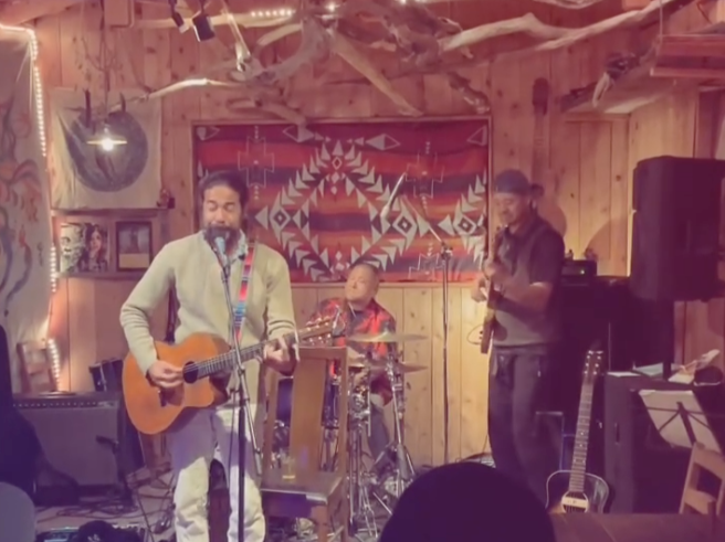 木の壁と装飾の中で、一組の男性と女性がギターを演奏しているライブ音楽の場面。