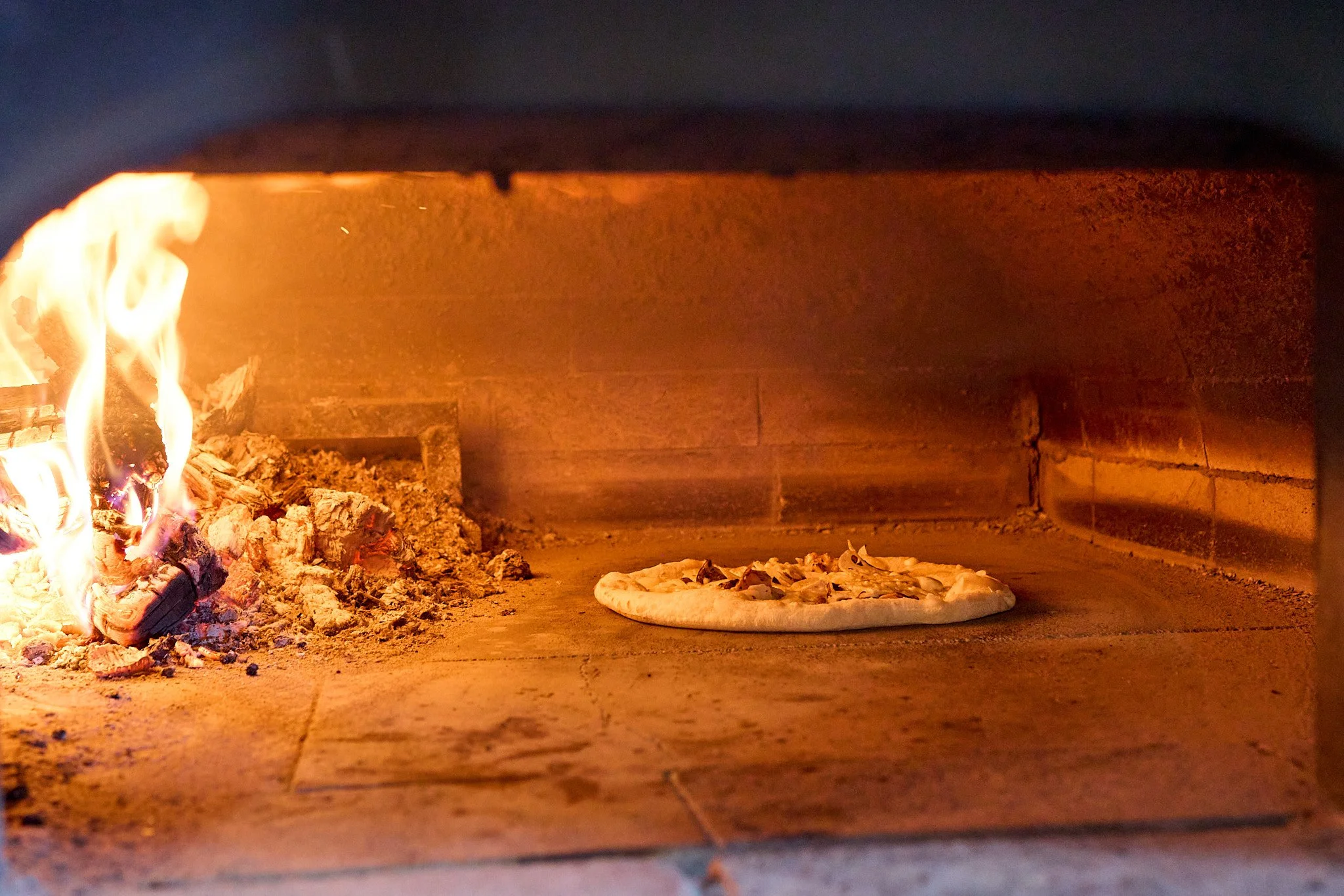 ピザがオーブンの中の床に置かれており、火が燃えているレンガの壁のオーブン内の様子
