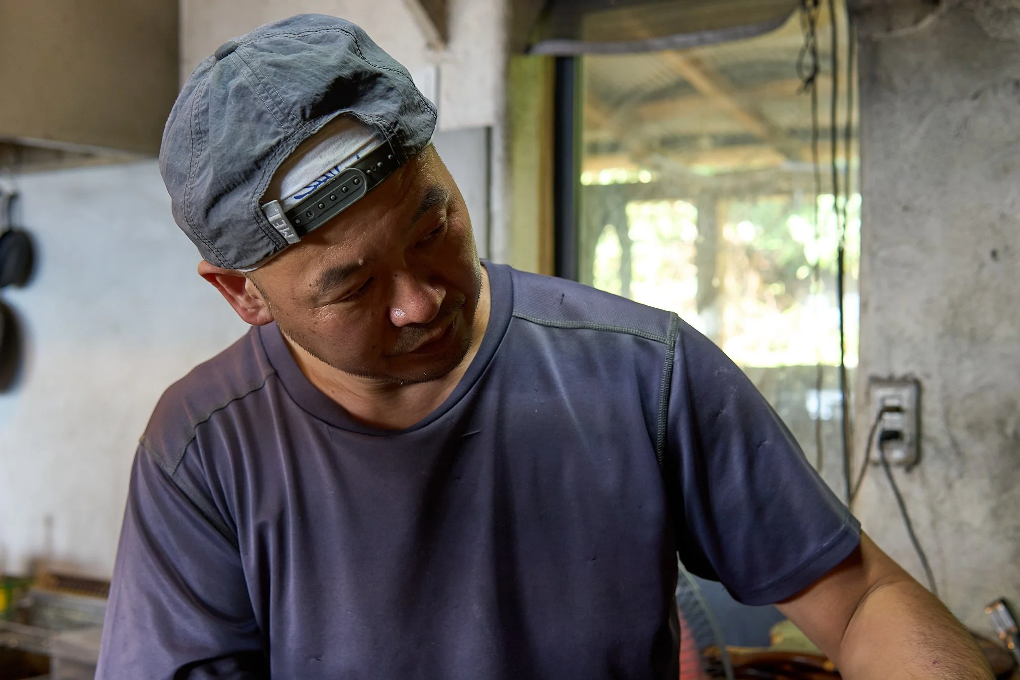 男性が作業をしている様子。灰色のキャップと濃紺のTシャツを着ている。室内の背景には窓と壁のひび割れが見える。