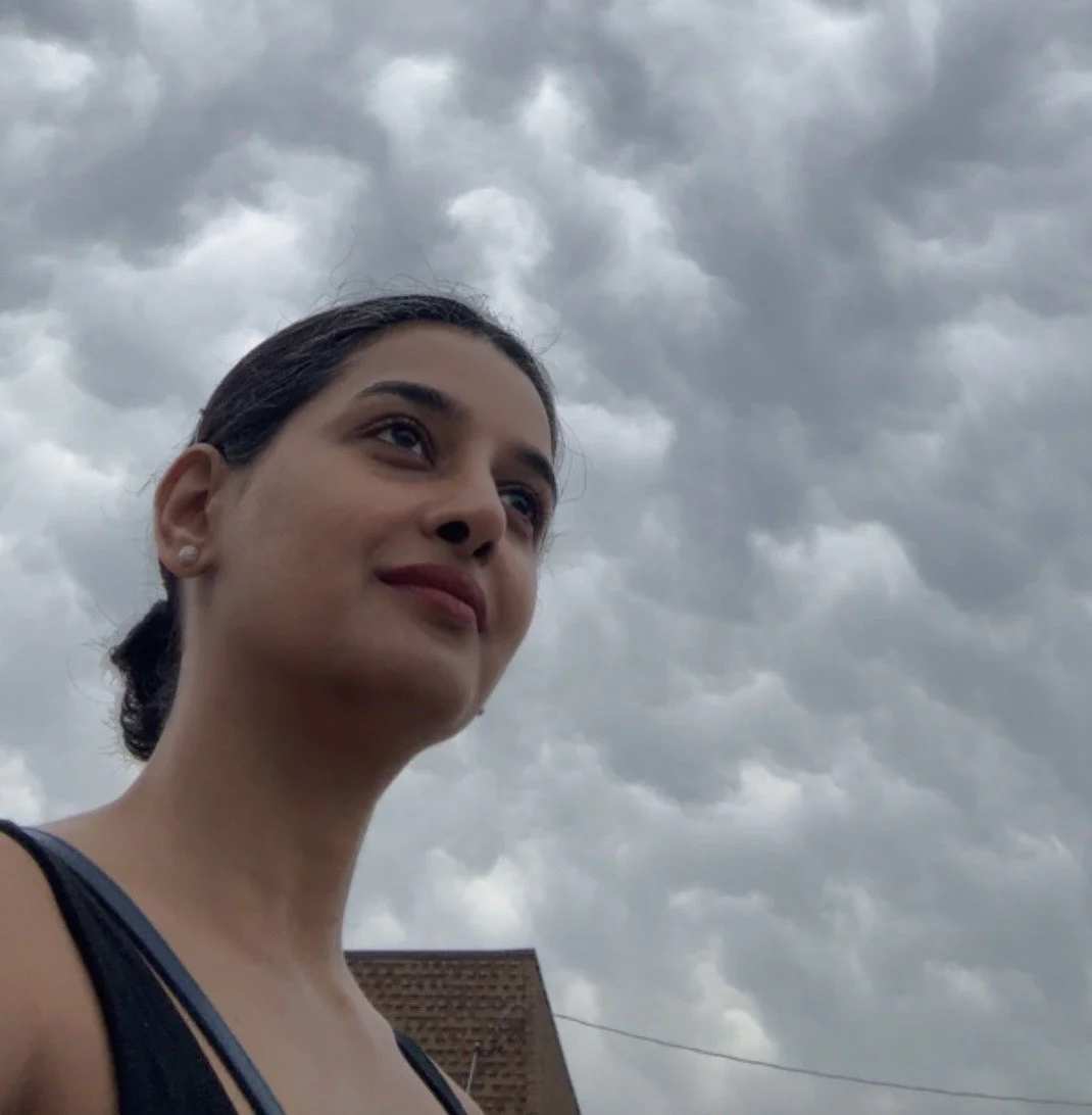 Young woman with dark hair pulled back, smiling slightly, wearing a sleeveless top, standing outdoors under cloudy skies.