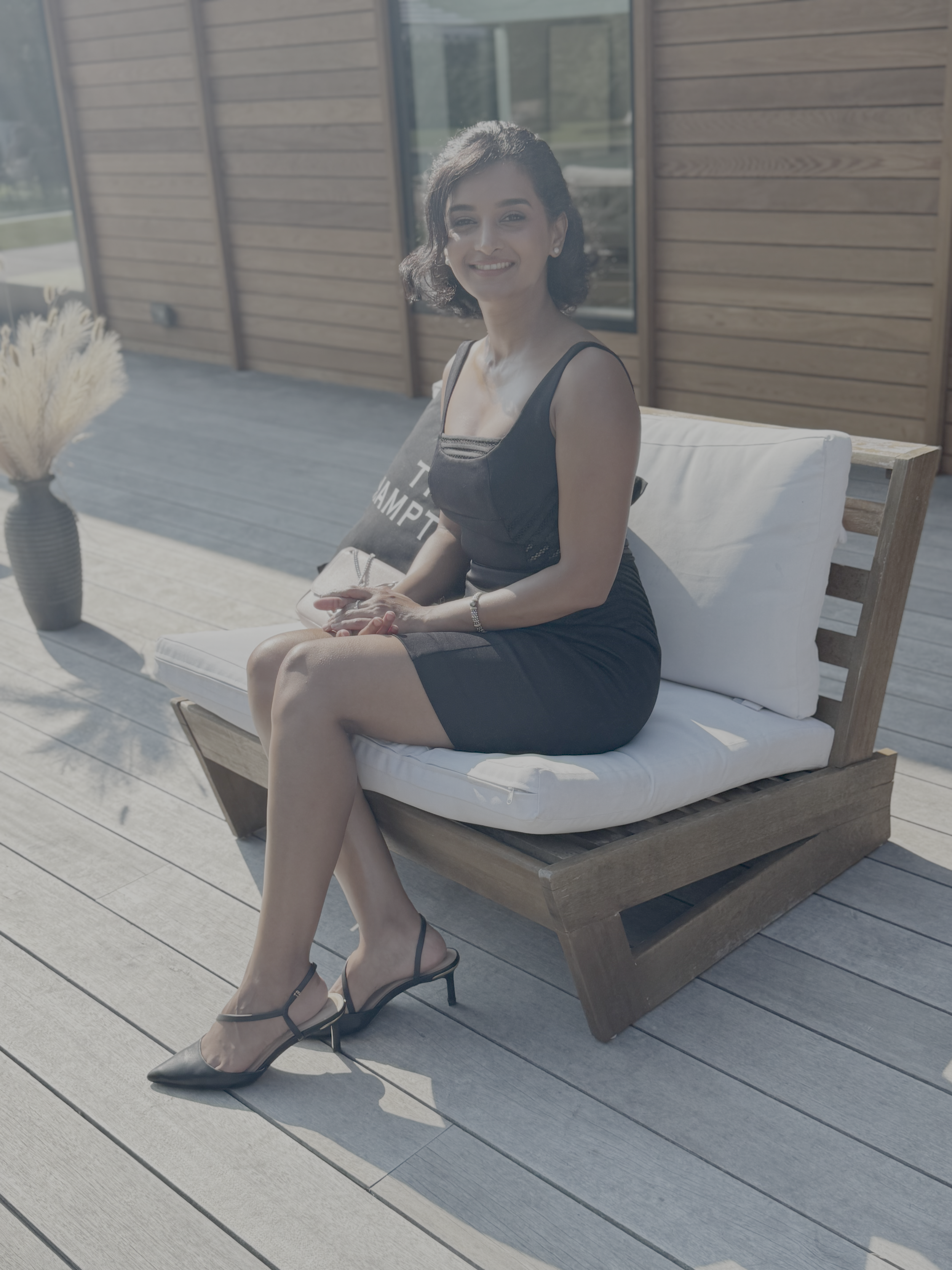 A woman sitting on a wooden outdoor bench on a patio, wearing a black dress and black high-heeled shoes, smiling at the camera.
