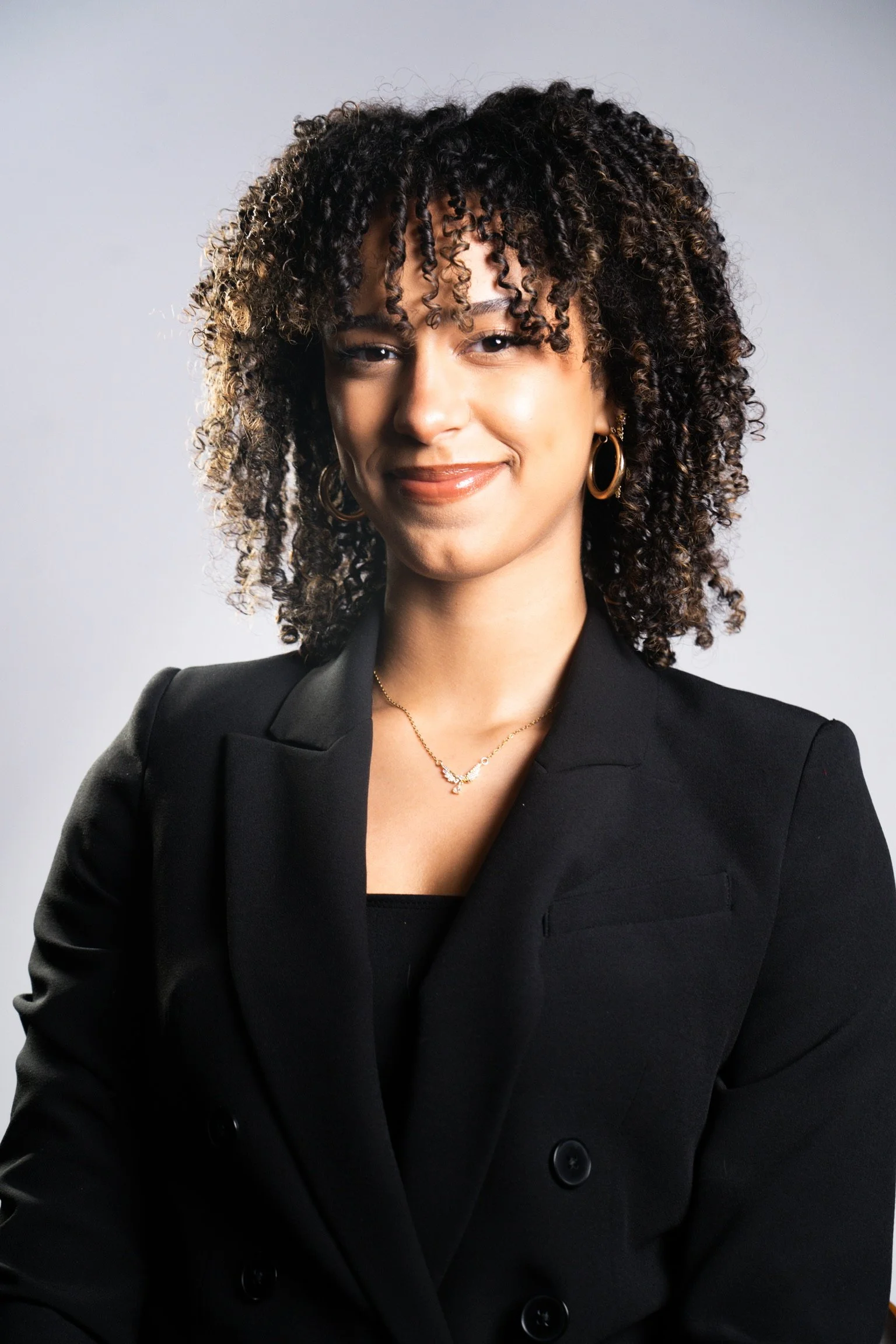 Portrait of a woman with curly hair wearing a black blazer, hoop earrings, a necklace, and smiling with a neutral background.