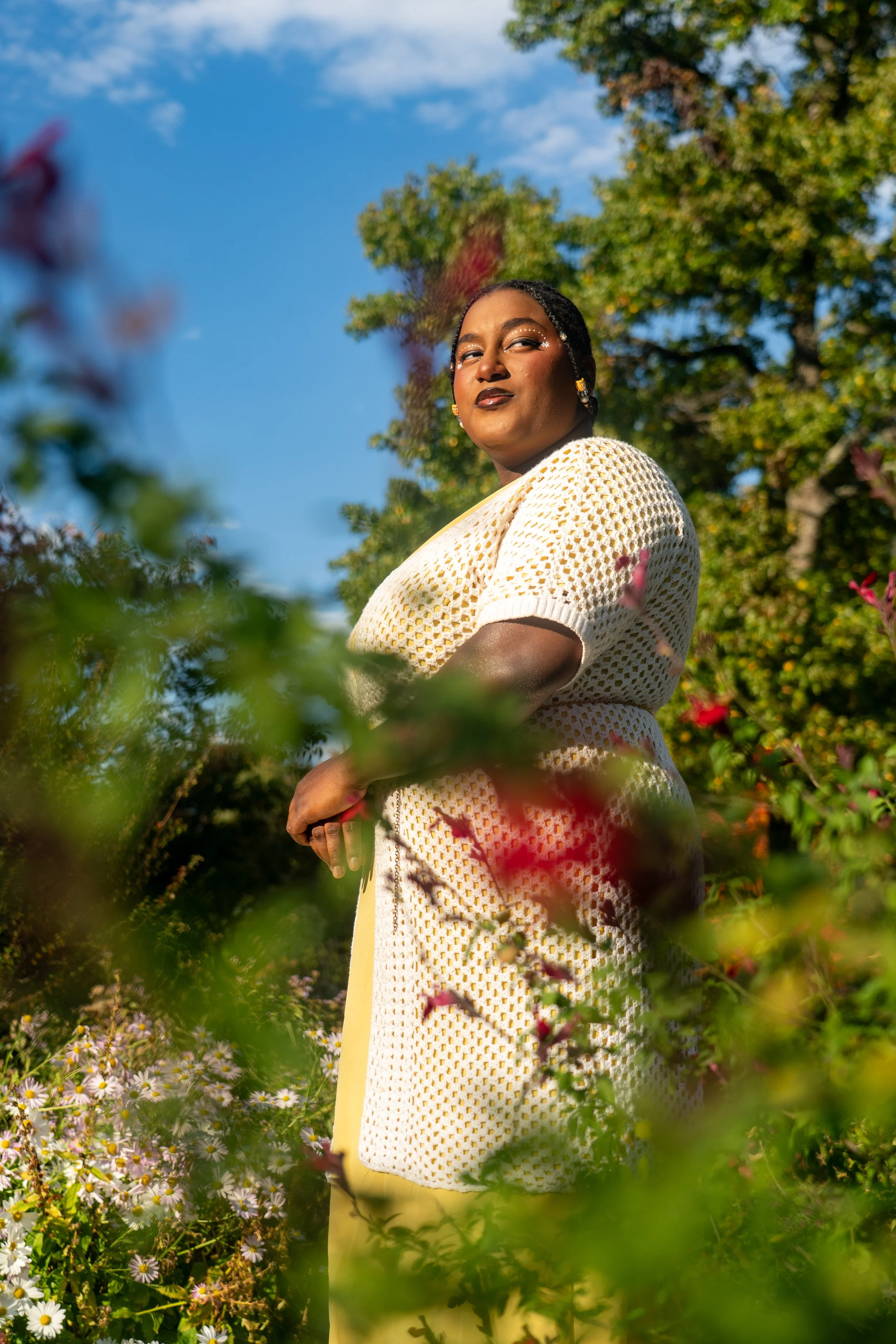 A woman standing outdoors among flowers and greenery, wearing a cream, open-knit dress, with dark lipstick and jewelry, looking confidently at the camera against a blue sky with scattered clouds.