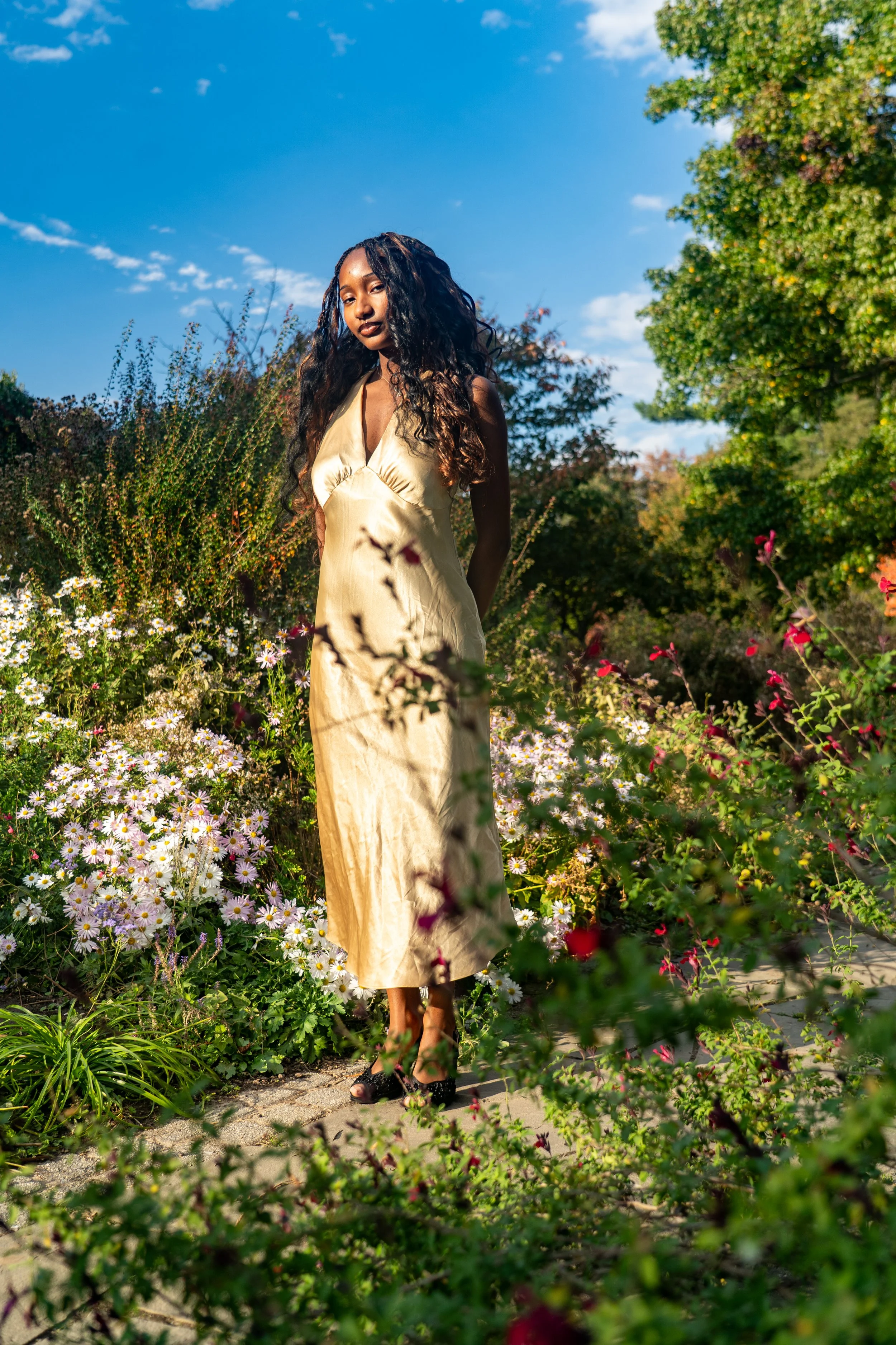 A woman in a gold dress standing in a flower garden under a blue sky with trees in the background.