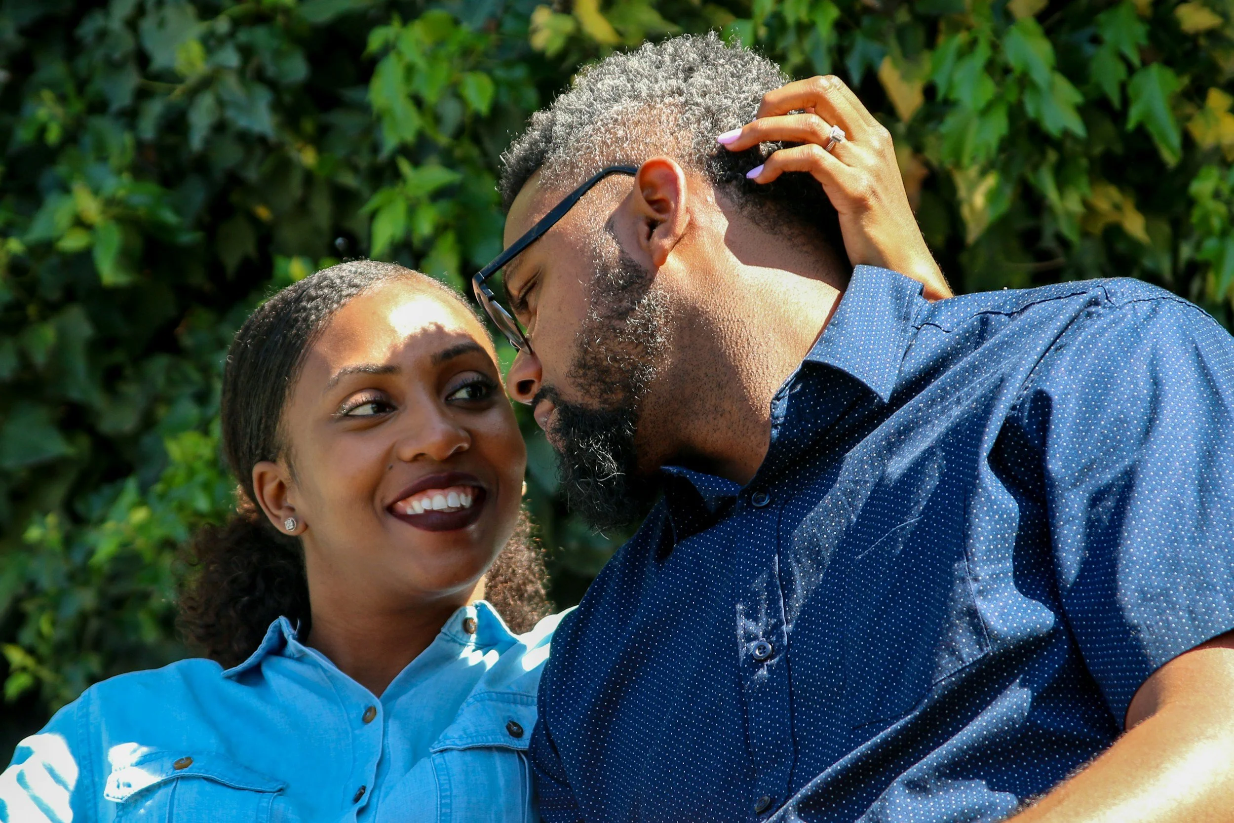 A smiling African American woman in a light blue shirt looking into the eyes of an older African American man with gray hair and beard, wearing glasses and a dark blue patterned shirt, with a background of green foliage.