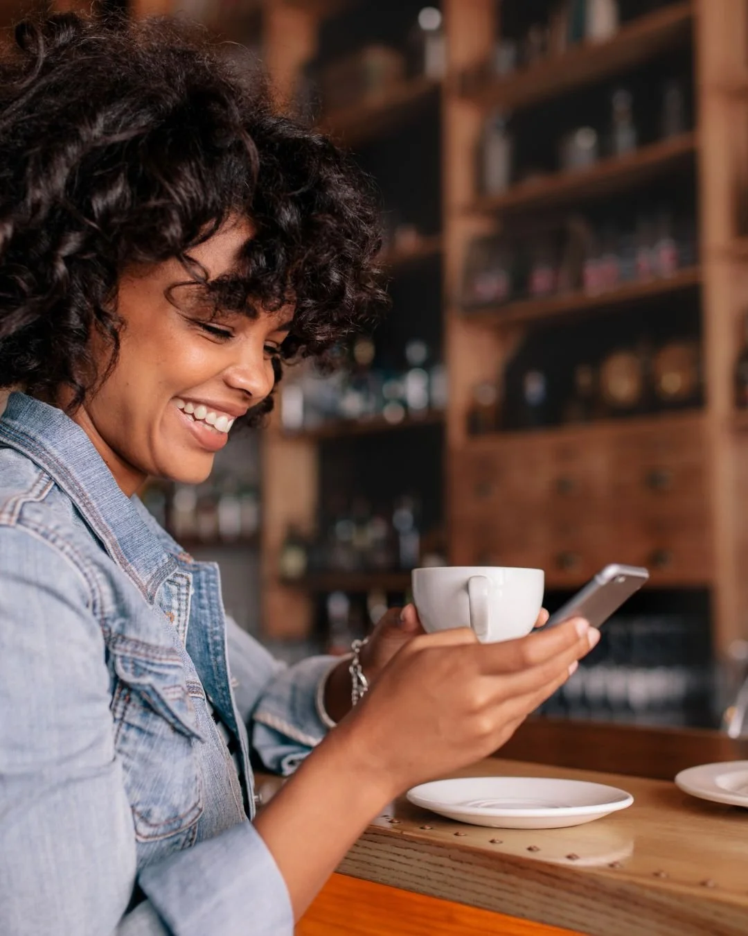 A woman sitting at a desk, smiling, looking at her smartphone, with a laptop and notepad in front of her.