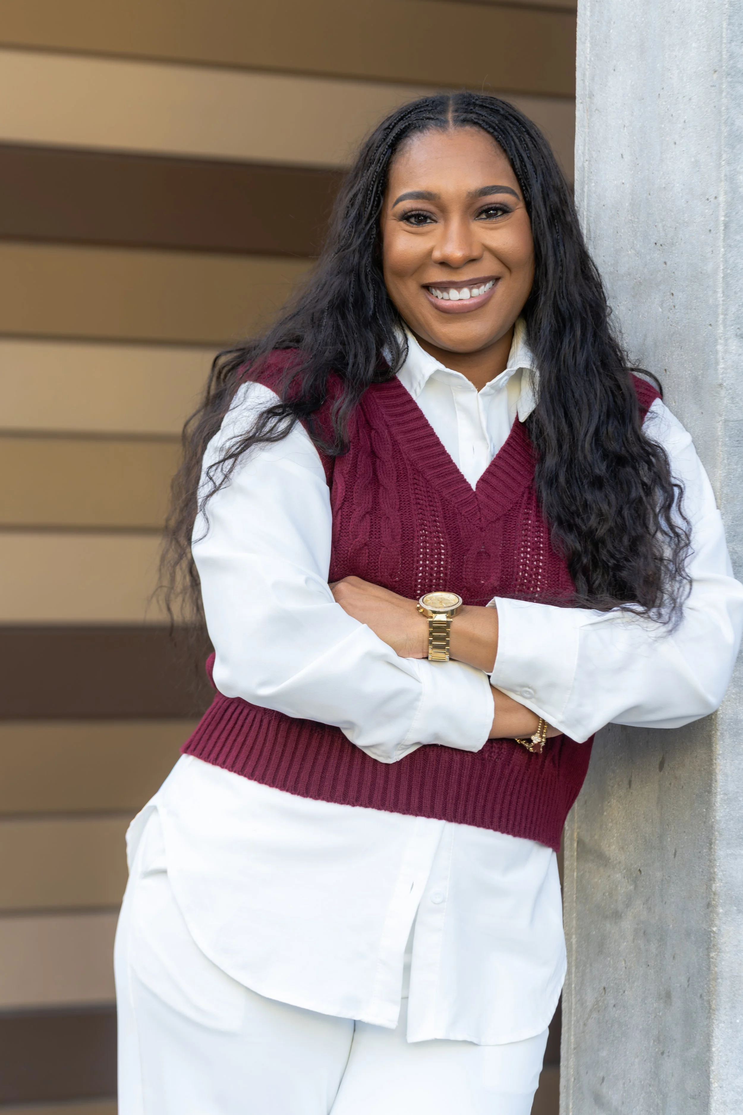 A woman with long curly black hair, wearing a white shirt, maroon sweater vest, and white pants, standing with crossed arms next to a concrete pillar, smiling at the camera.