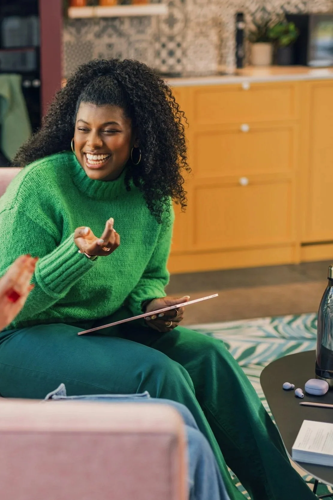 A woman with curly black hair, wearing a bright green sweater, smiling and gesturing while sitting at a table in a cozy, modern living room.