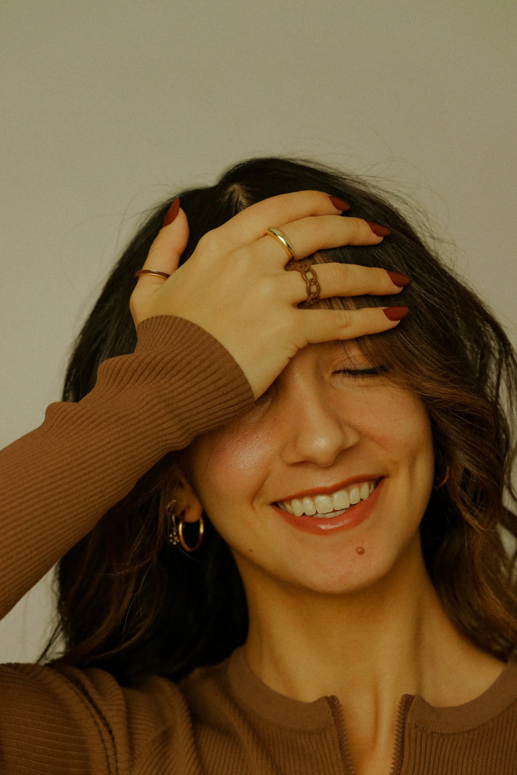 A woman with dark wavy hair, earrings, and rings on her fingers is smiling with her eyes closed, wearing a brown top, with her hand on her forehead.