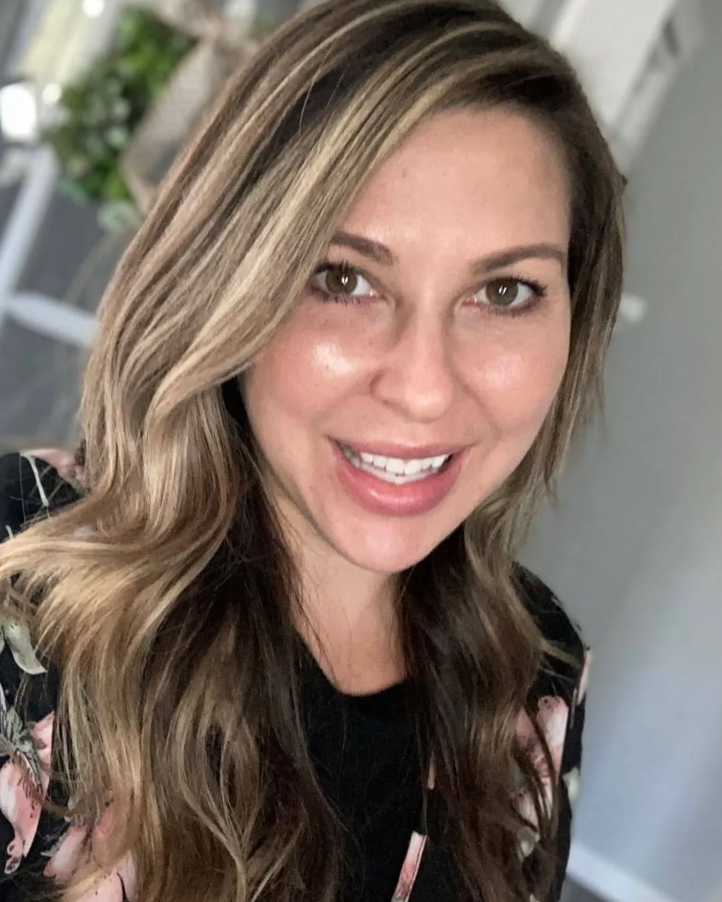 A smiling woman with long, wavy, light brown hair with blonde highlights, wearing a black top with floral print, indoors with a grey wall and potted plant in the background.