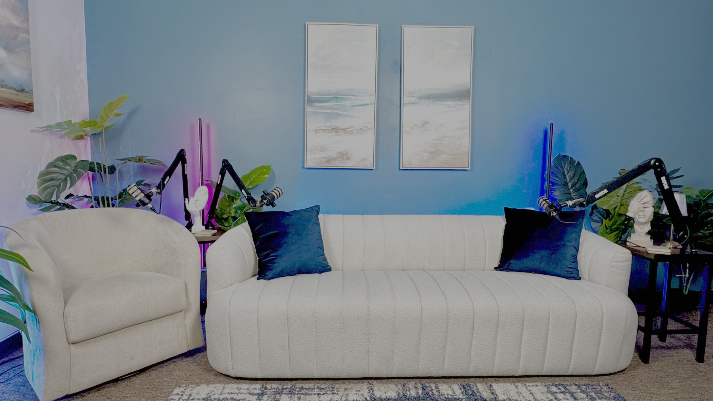 Living room with beige armchair and white sofa with dark blue pillows, blue painted wall with two landscape paintings, plants, and two black side tables with microphones and decorative busts, illuminated with purple and blue accent lights.