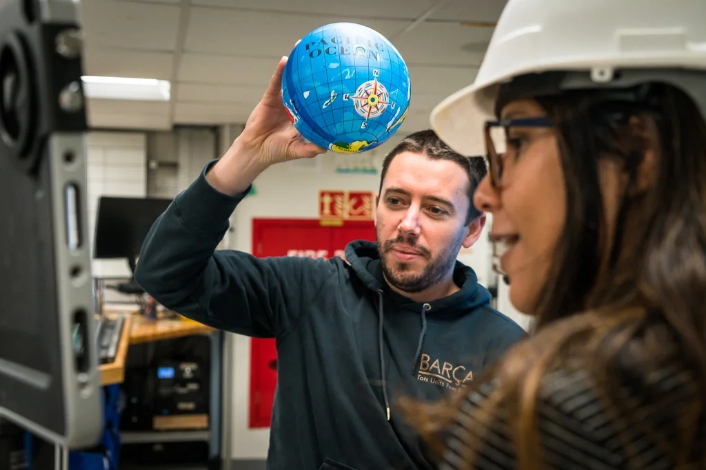 Dr Lloyd White holding a small globe next to a woman wearing safety glasses and a hard hat, in an indoor setting with equipment in the background. This photo was taken by Dr Vivien Cumming during International Ocean Discovery Program Expedition 369.
